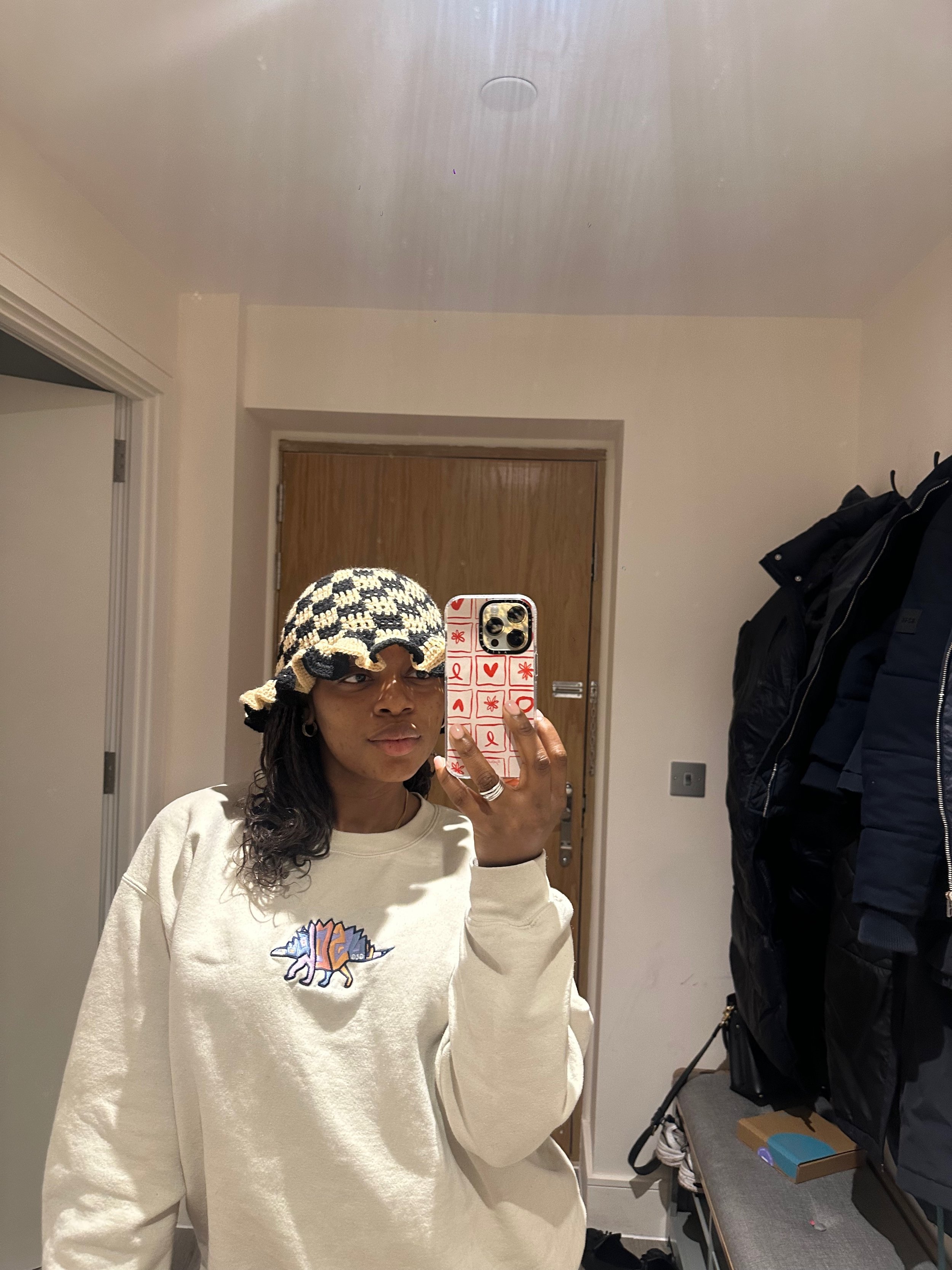 Person taking a mirror selfie in a room with a king hat. The person wears a beige sweatshirt with a colorful logo. A wooden door and a coat rack with black jackets are visible in the background.