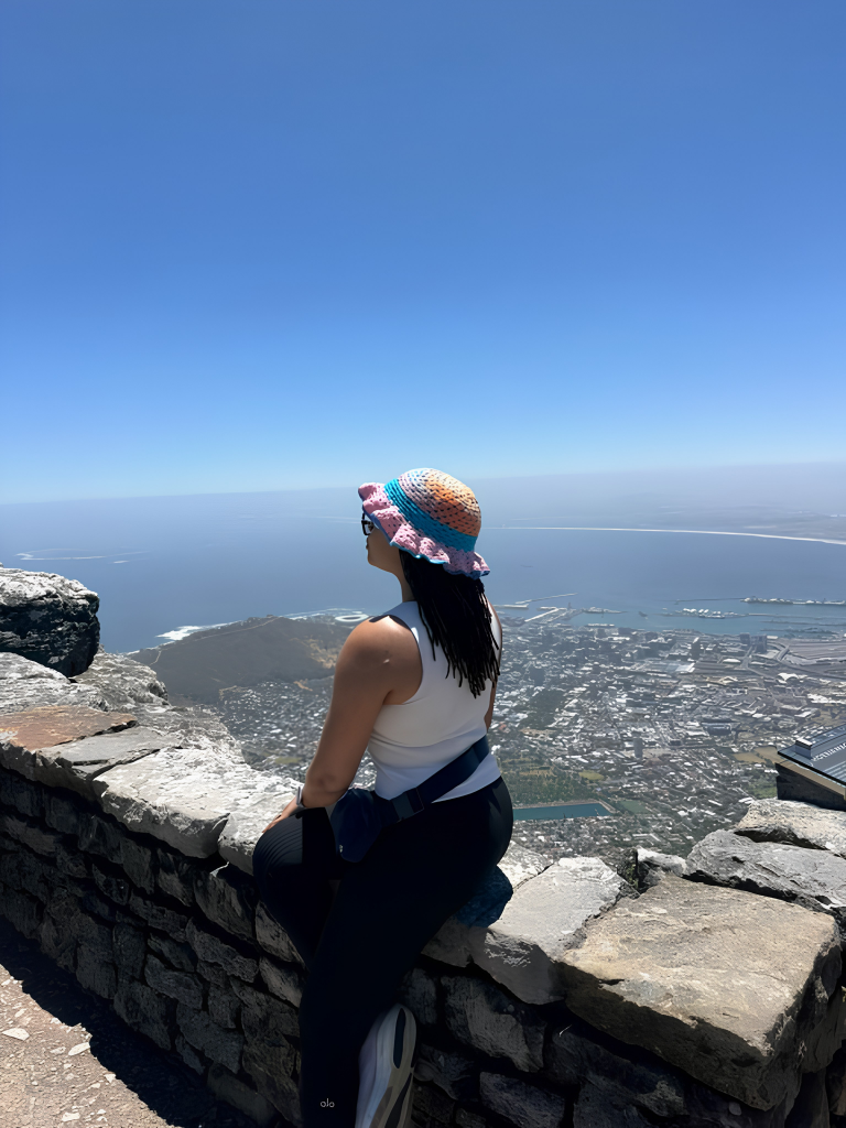 A woman sitting on a stone ledge on a mountain, overlooking a coastal town and the ocean under a clear blue sky.