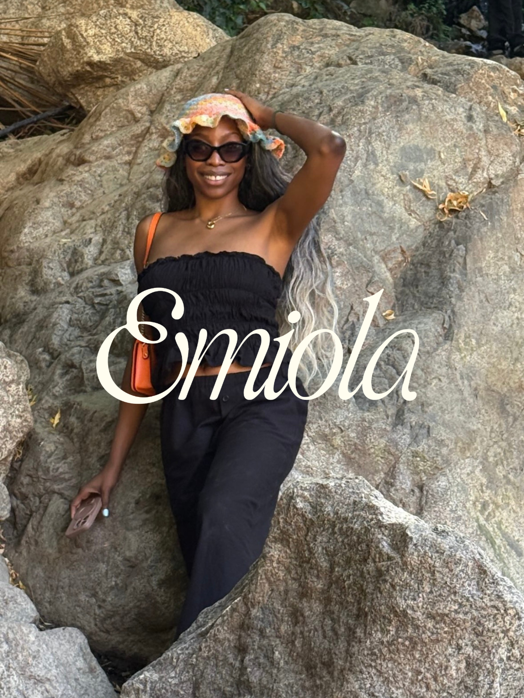 A woman standing among large rocks outdoors, smiling, wearing sunglasses, a floral hat, a black strapless top, and black pants, carrying an orange purse.