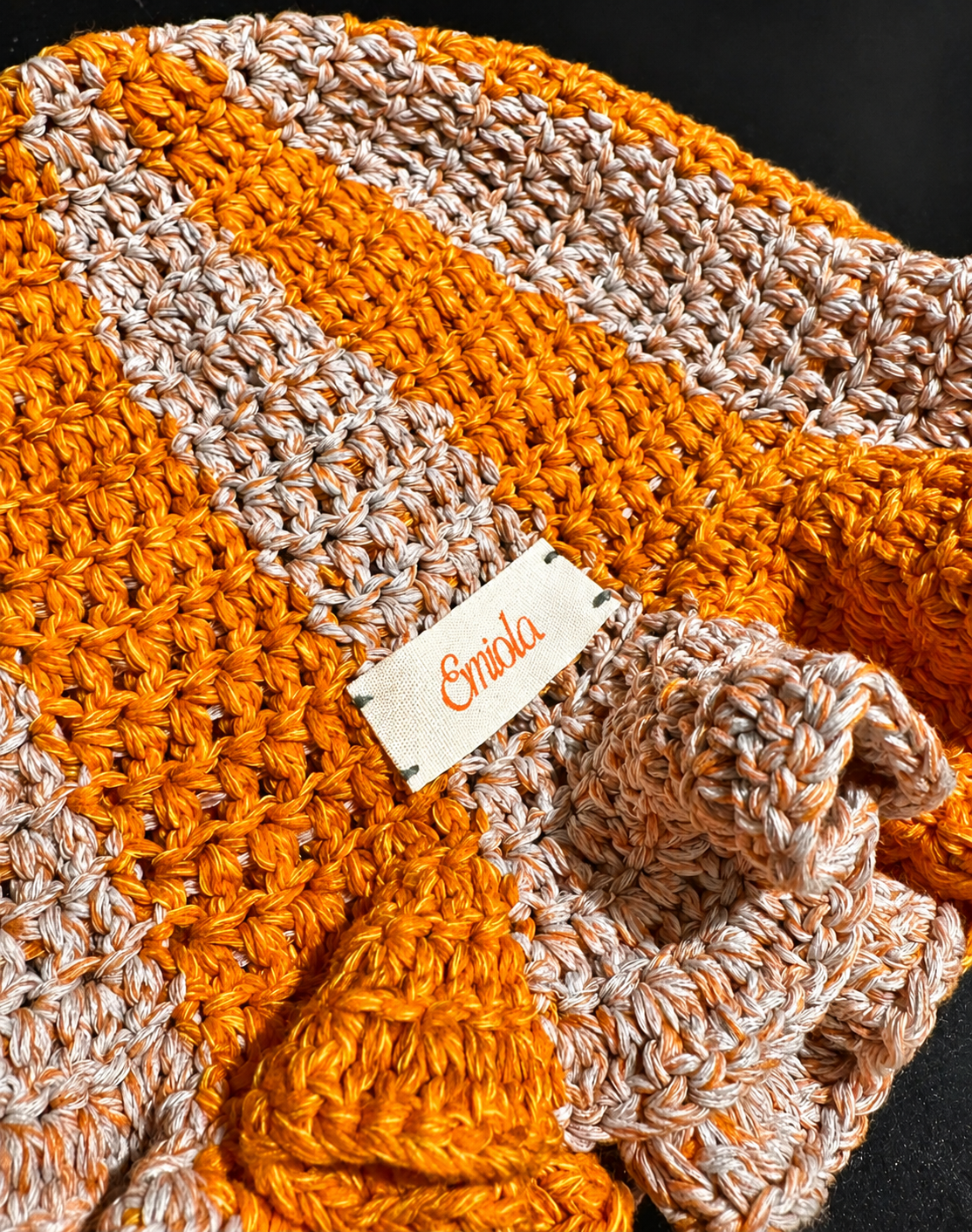 Close-up of a hand-crocheted blanket with orange and cream yarn, featuring a small fabric label with orange text reading 'Emilda'.