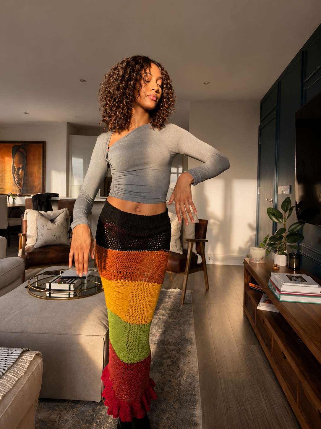 A woman with curly hair standing in a living room, wearing a gray long-sleeve crop top and a colorful crochet skirt, with sunlight streaming in.