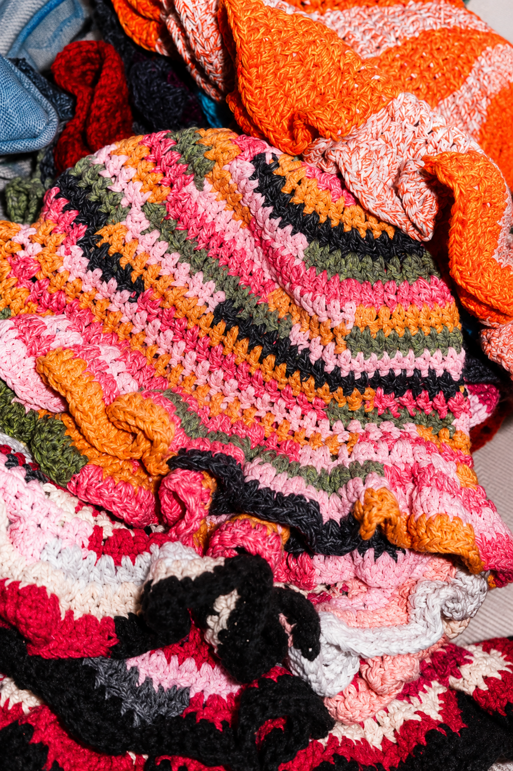 A pile of colorful, crocheted blankets with various patterns and stripes in bright colors including pink, orange, black, white, and green.