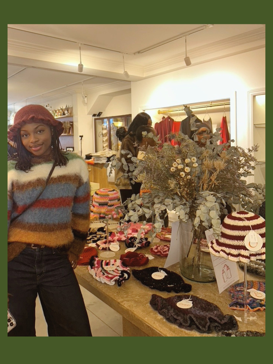 The Founder of ByEmiola with a brown and blue striped sweater and a red knit hat stands in a boutique store with fall-themed accessories and decorations on display, including hand-knit hats, scrunchies, and a large floral arrangement.