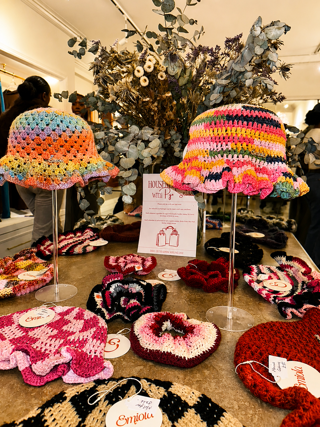 Display of colorful crochet hats and accessories on a table with a large bouquet of dried flowers in the background at an indoor craft or holiday market.