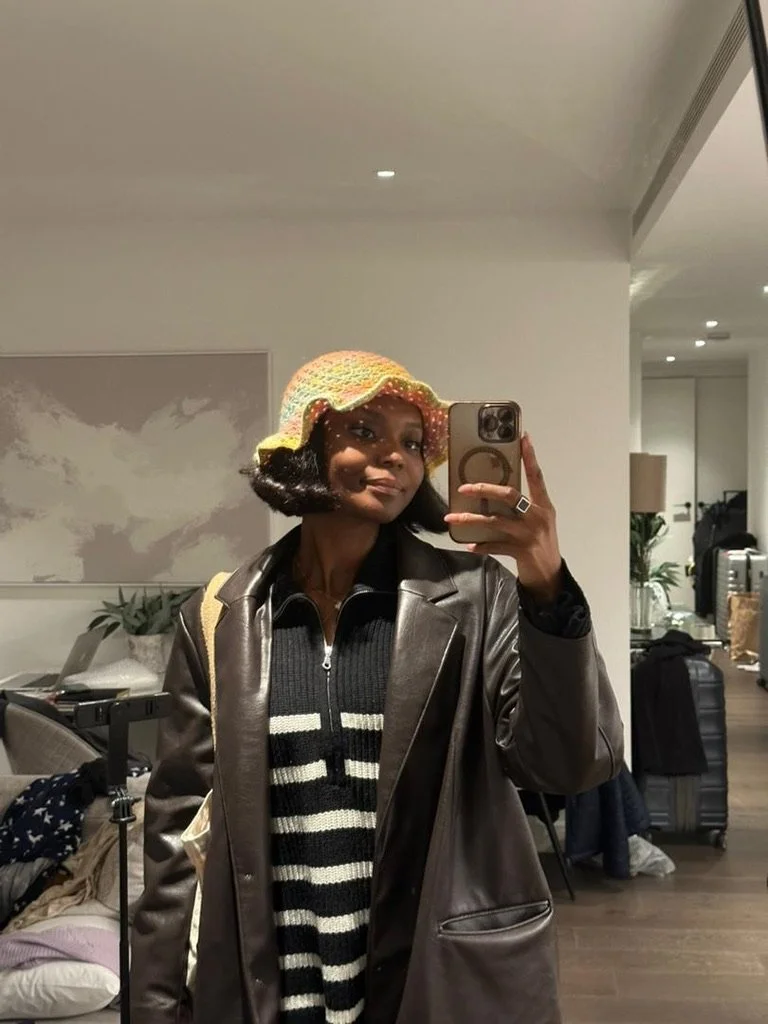 A woman taking a mirror selfie, wearing a colorful knitted hat, a black and white striped sweater, and a brown leather jacket, in what appears to be a hotel room or modern living space.