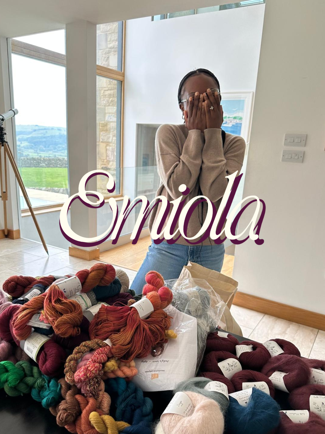 A woman covers her face with her hands, standing behind a table with various skeins of colorful yarn, in a room with large windows and a scenic view outside.