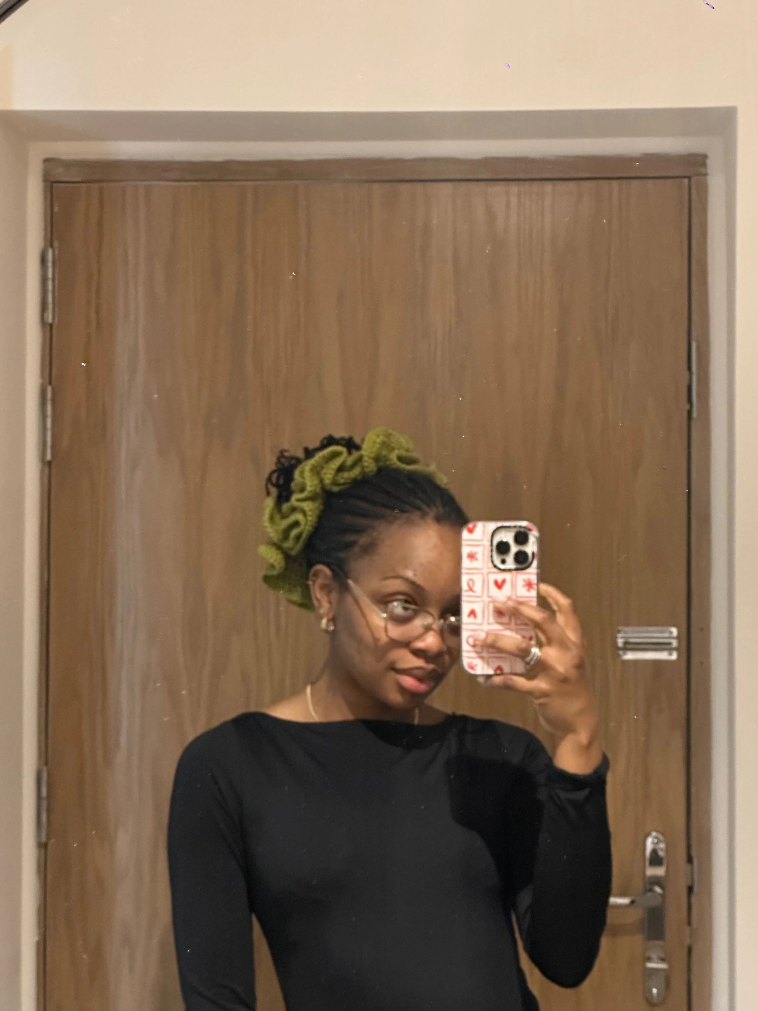 A woman taking a mirror selfie in front of a wooden door with a towel on her head, wearing glasses and a black top.