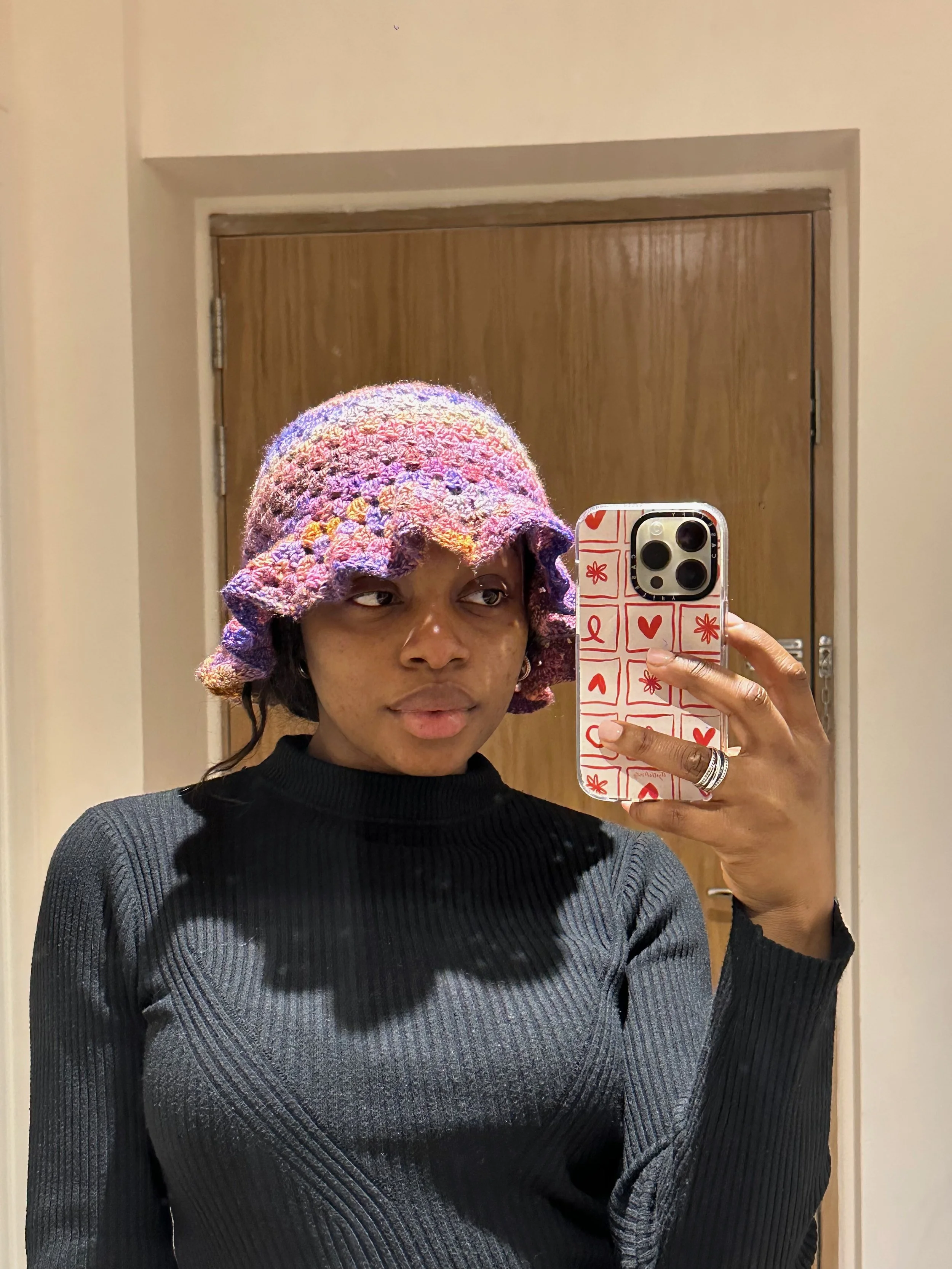 A person taking a mirror selfie with a smartphone, wearing a colorful crocheted hat and a black and gray striped sweater in a room with a wooden door in the background.
