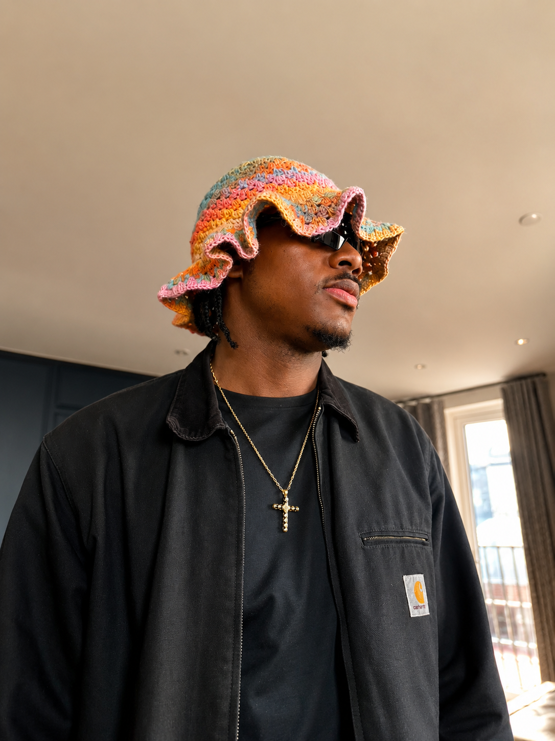 A man wearing a colorful, ruffled crochet hat with pink, yellow, orange, and blue colors, black sunglasses, a black Carhartt jacket, a black shirt, and a gold cross necklace standing indoors near a window.