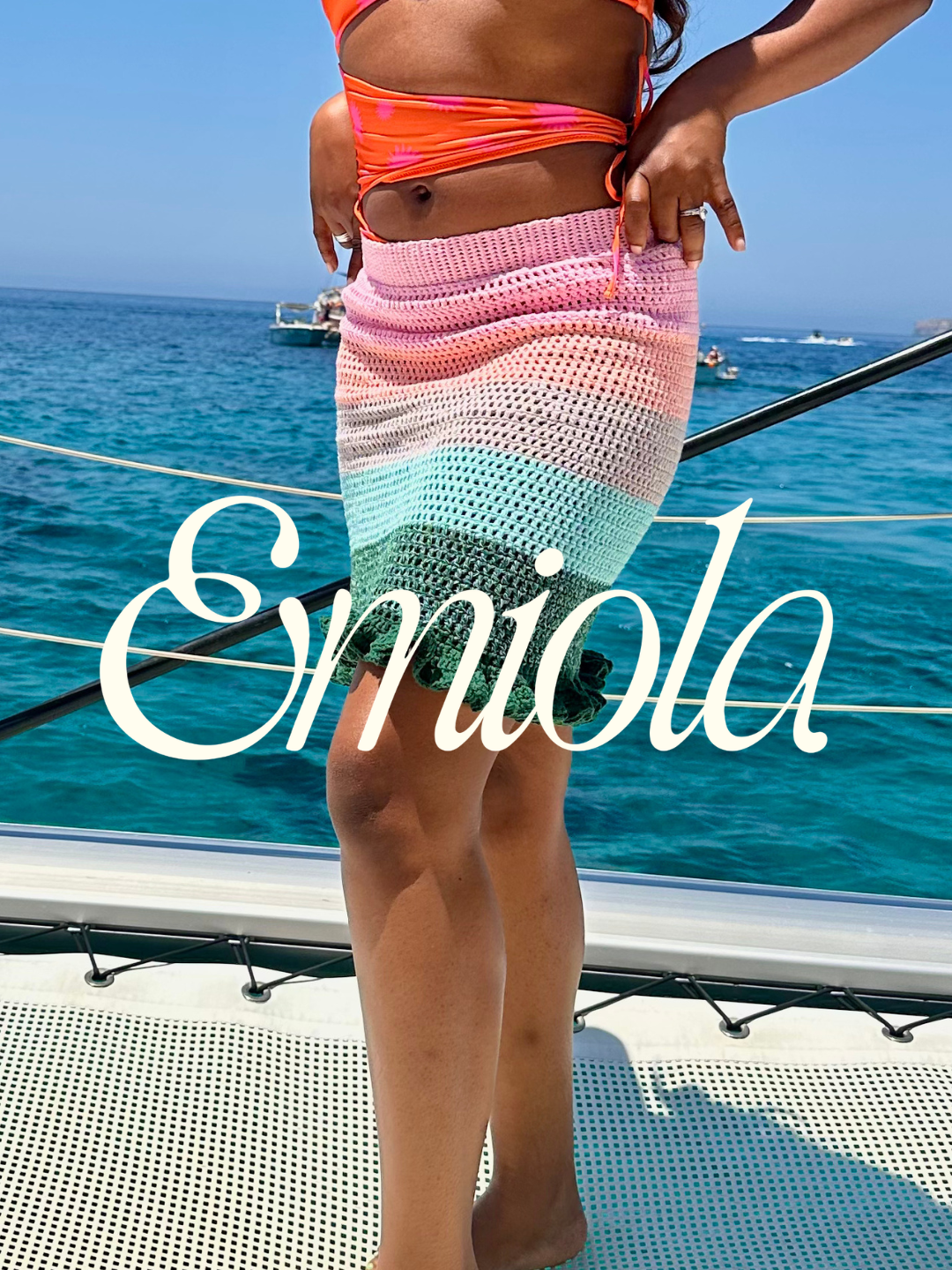 A woman in colorful swimwear standing on a boat with ocean in the background, with the word 'Emiola' written across the image.