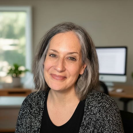A woman with shoulder-length gray hair smiling at the camera, sitting in an office with a computer and a window in the background.