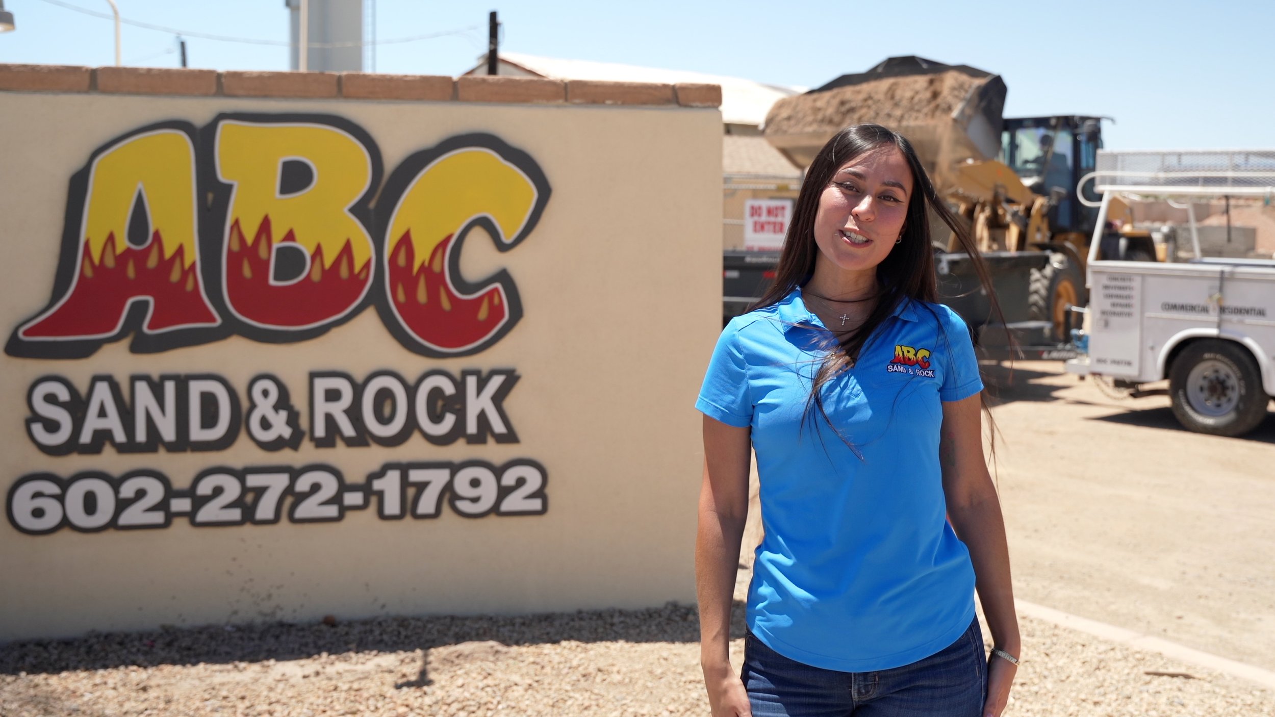 A young woman with long dark hair wearing a blue polo shirt with a logo, standing outdoors near a sign that reads "ABC Sand & Rock" with a phone number, and construction equipment in the background.