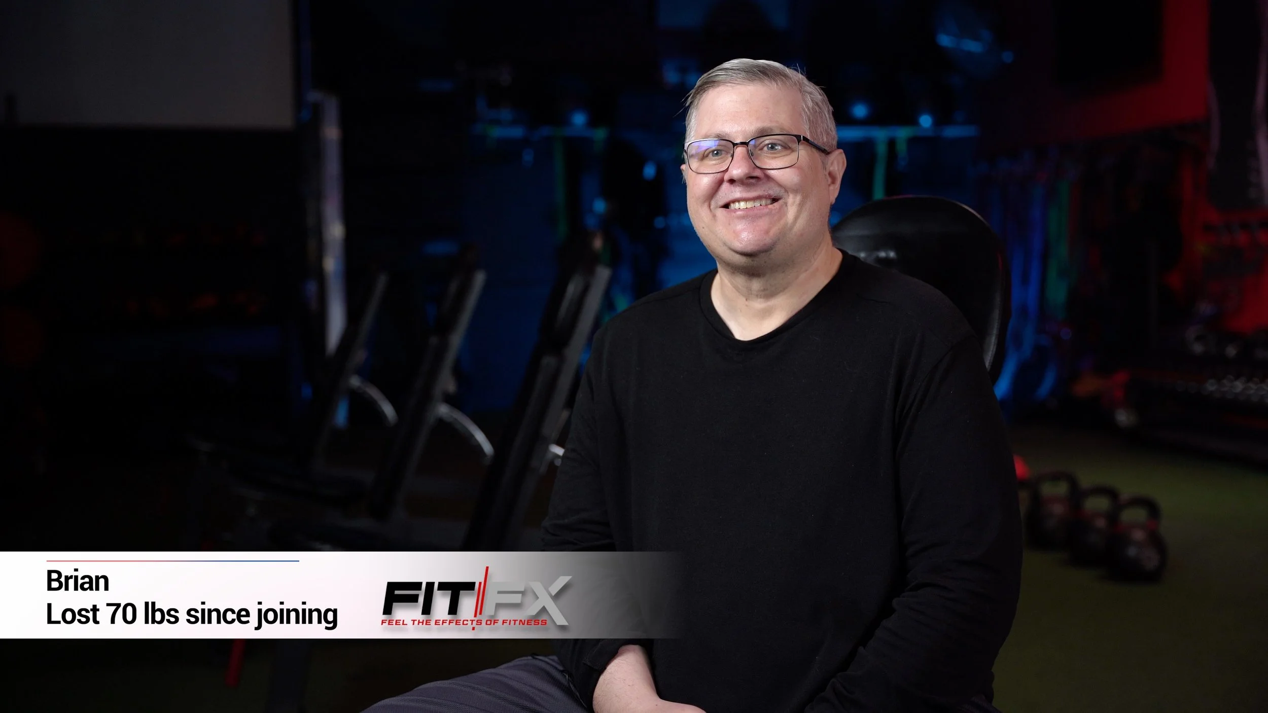 A man wearing glasses and a black shirt sitting in a fitness gym, smiling at the camera with exercise equipment in the background. giving a testimonial on the fitness studio he works out of in Gilbert AZ