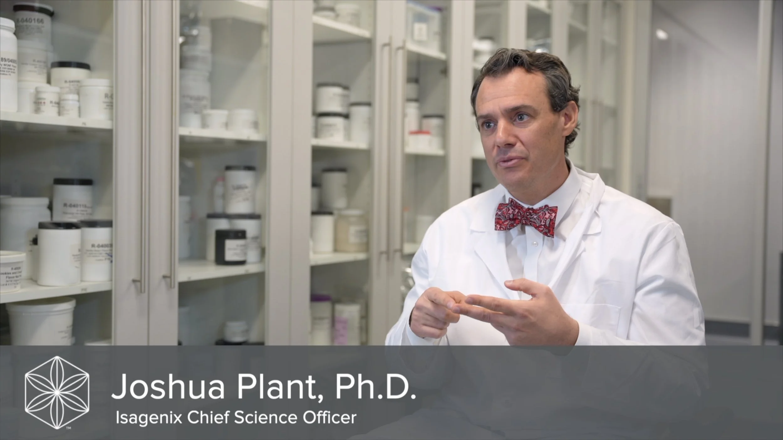 A man in a lab coat with a red patterned bow tie talking in a laboratory with shelves of labeled bottles and containers in the background. The overlay text reads "Joshua Plant, Ph.D., Isagenix Chief Science Officer."