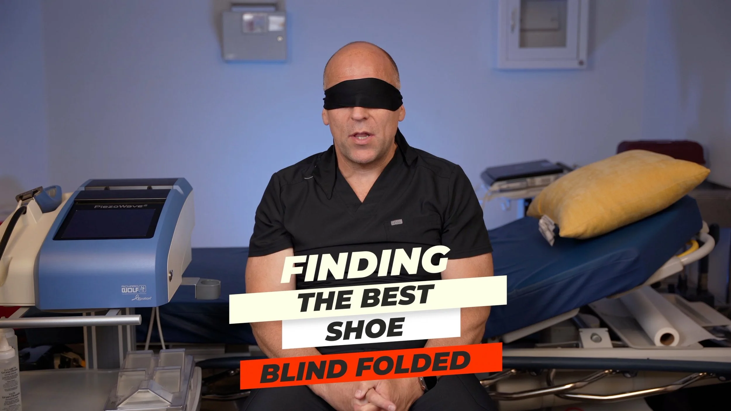 A man in black medical scrubs sitting on a hospital bed with a yellow pillow and medical equipment in the background, blindfolded, with text overlay 'Finding the Best Shoe Blind Folded'.