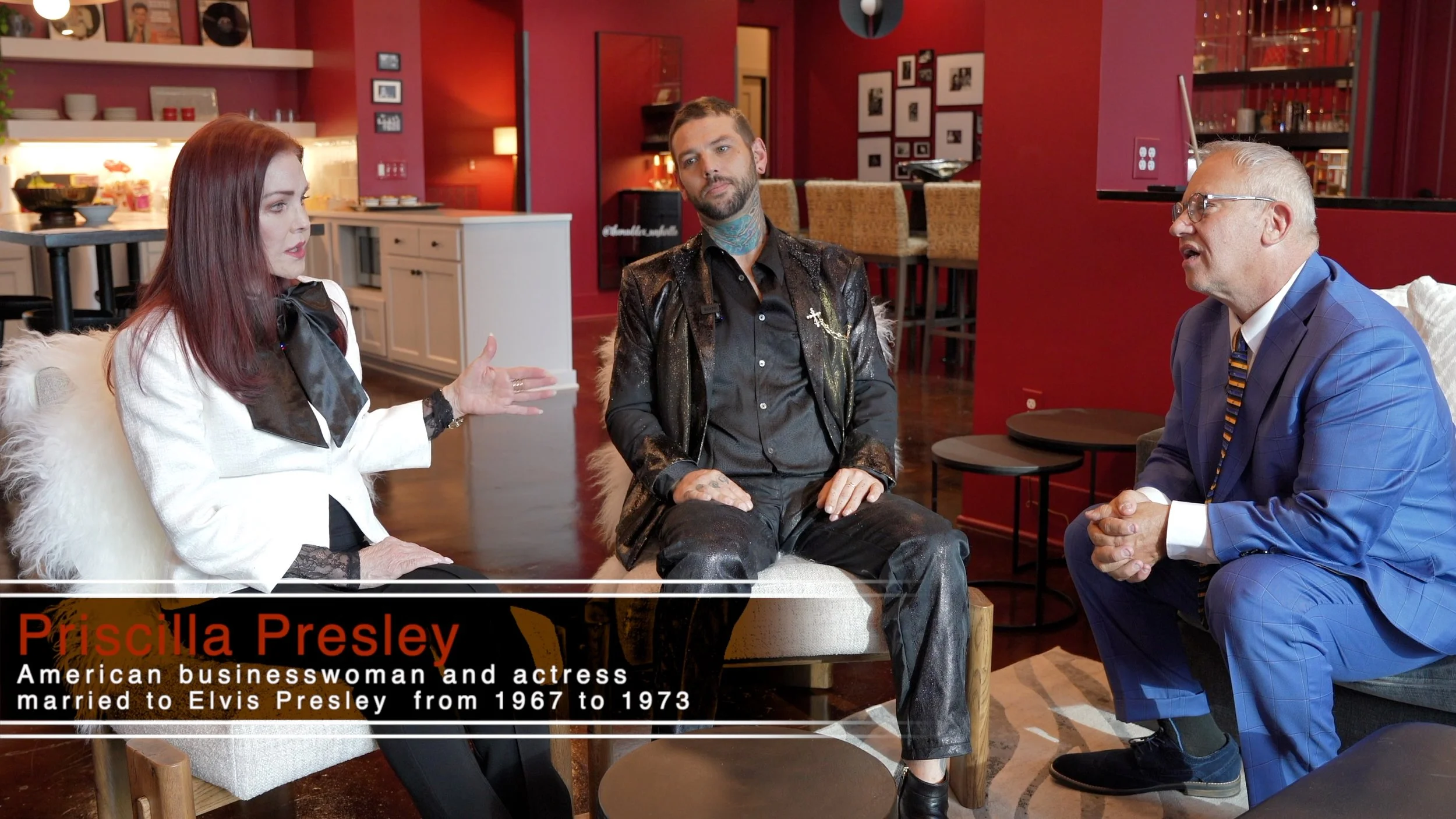 Priscilla Presley, an American businesswoman and actress, sitting on a chair in a conversation with two men in a well-lit, modern room with red walls. She is wearing a white blazer with black lace details and a black bow at the neck. One man, in a black shiny jacket and black pants, is sitting with his hands on his lap, and the other man, in a blue checkered suit, is seated with his hands clasped, listening attentively. There is a caption overlay with her name and brief biography.