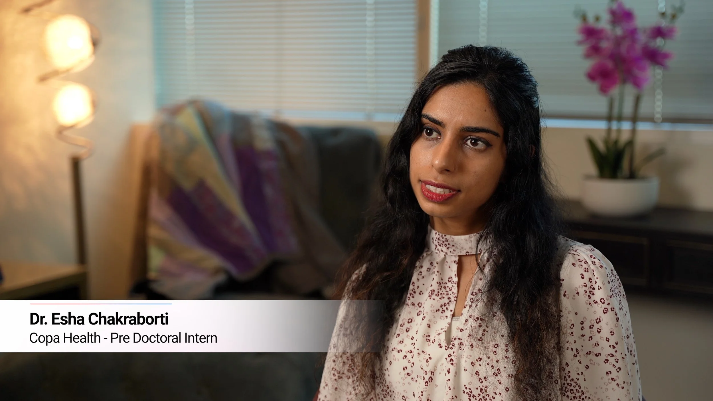 A young woman with long, dark, wavy hair, wearing a patterned blouse, sitting in a room with neutral decor and a potted orchid on a side table in the background in an interview giving her testimonial on working for copa health in mesa AZ