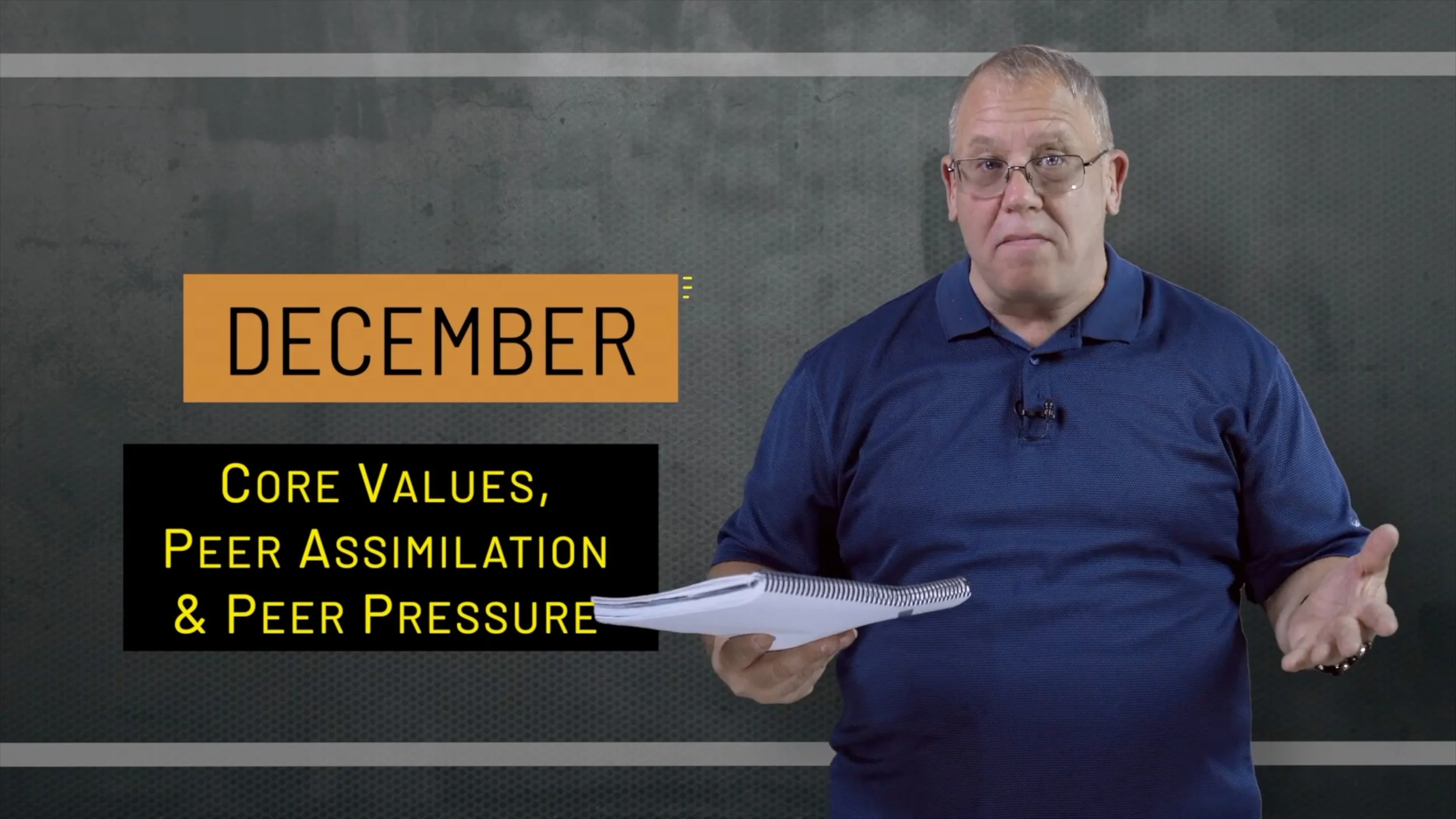 A man in a blue shirt holding papers presents a slide with the text "December" and "Core Values, Peer Assimilation & Peer Pressure" written on it, in front of a screen or backdrop.