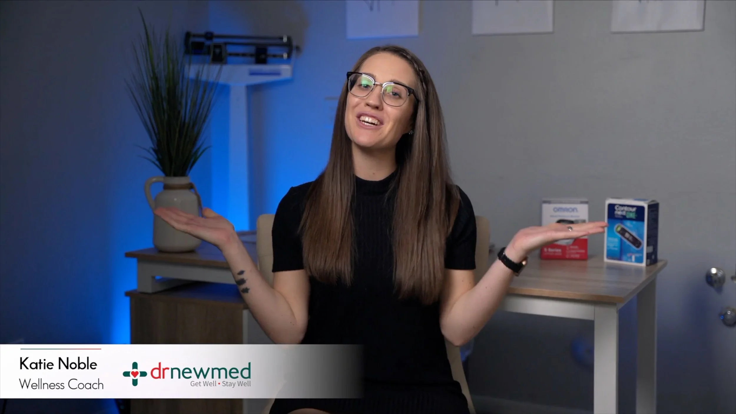 Wellness coach Katie Noble smiling and gesturing with her hands in a modern office setting, with wellness products on the table behind her.