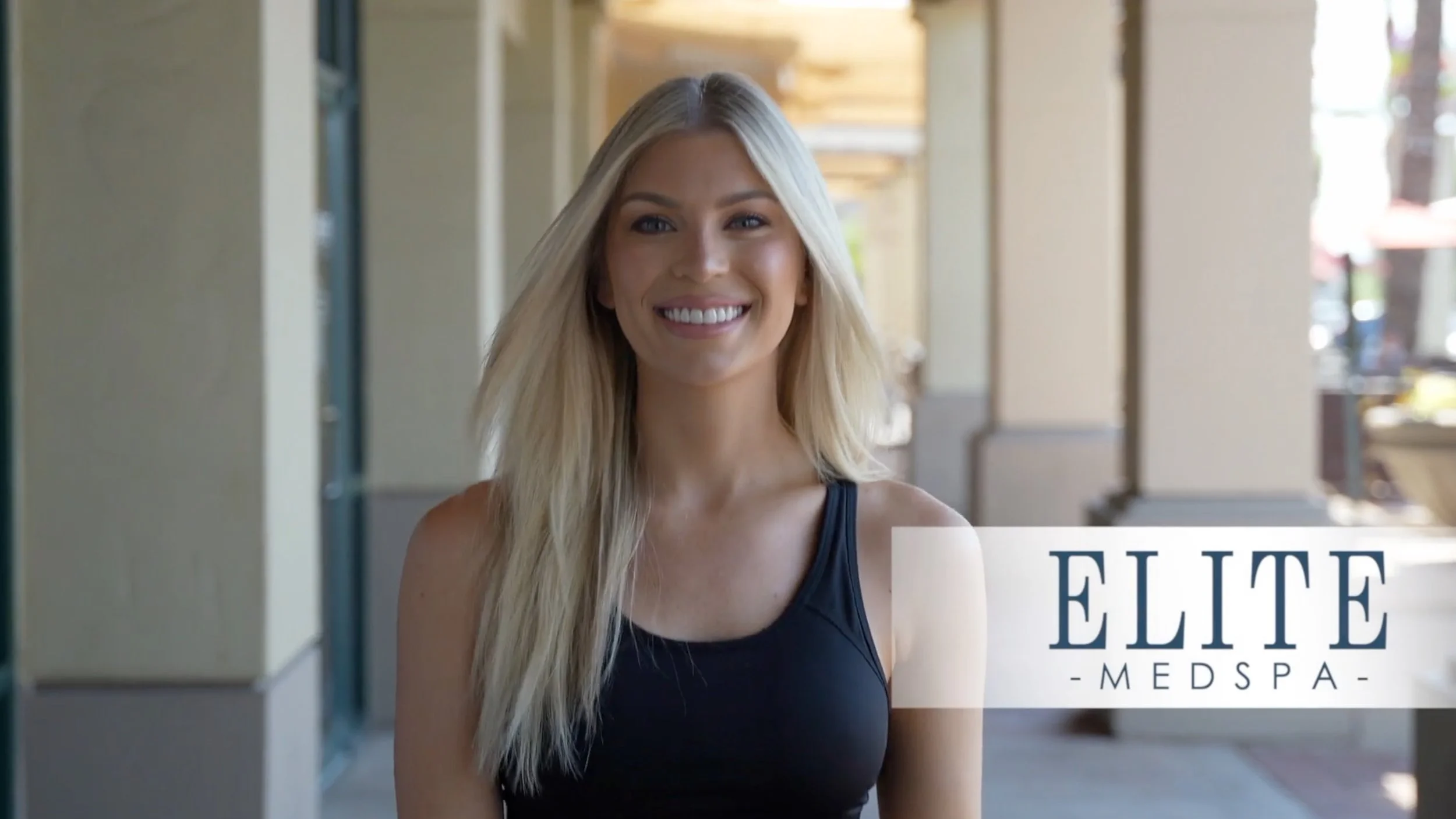 A young woman with long blonde hair smiling while walking outside in a covered area with columns, wearing a black tank top, with a logo saying ELITE MEDSPA in the lower right corner.