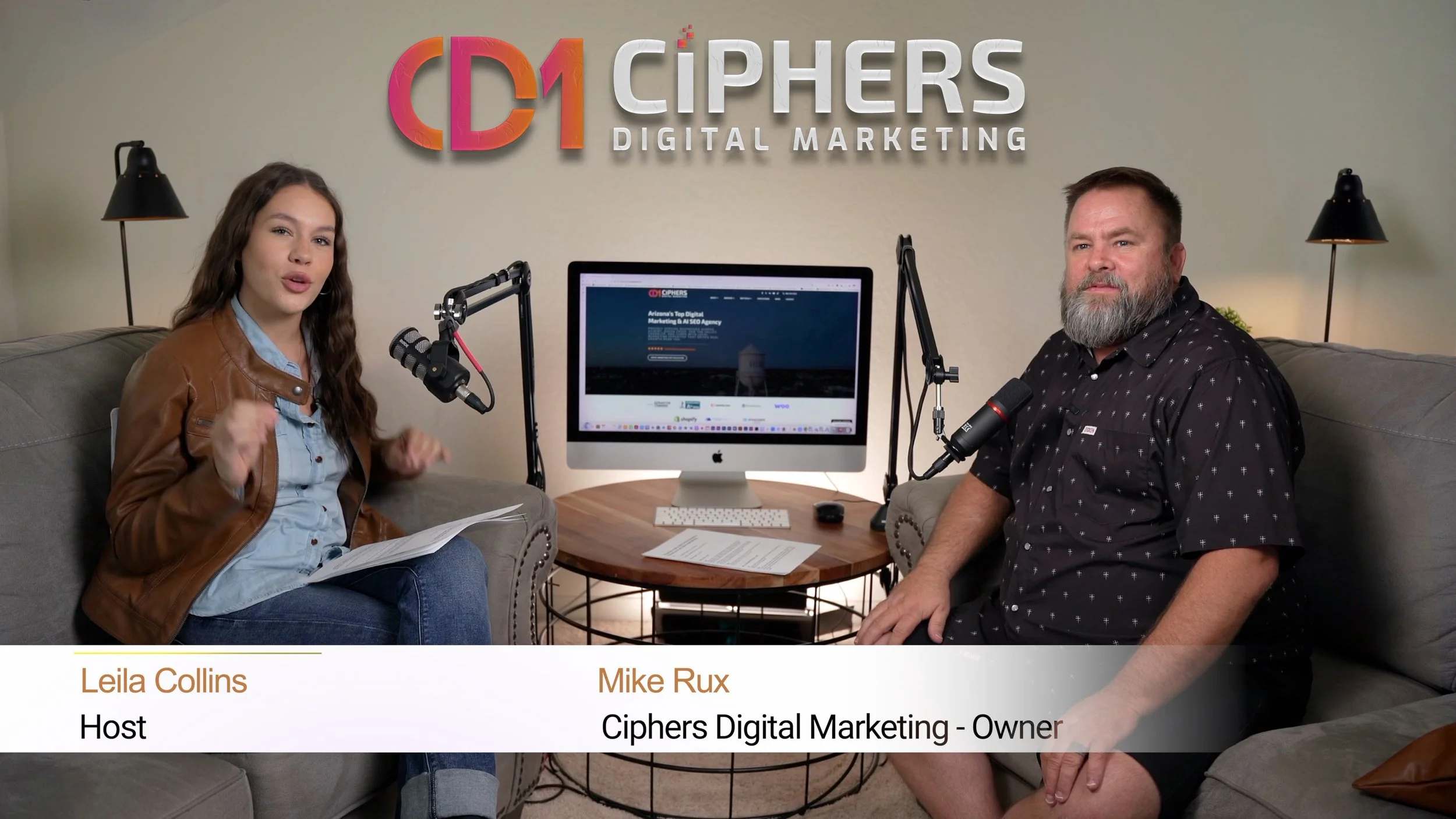 A woman and a man sit on sofas in a podcast studio with a sign that reads 'C1 CIPHERS DIGITAL MARKETING' behind them. The woman is speaking into a microphone and has a notebook on her lap, and the man is looking at the camera with a computer on a table between them.