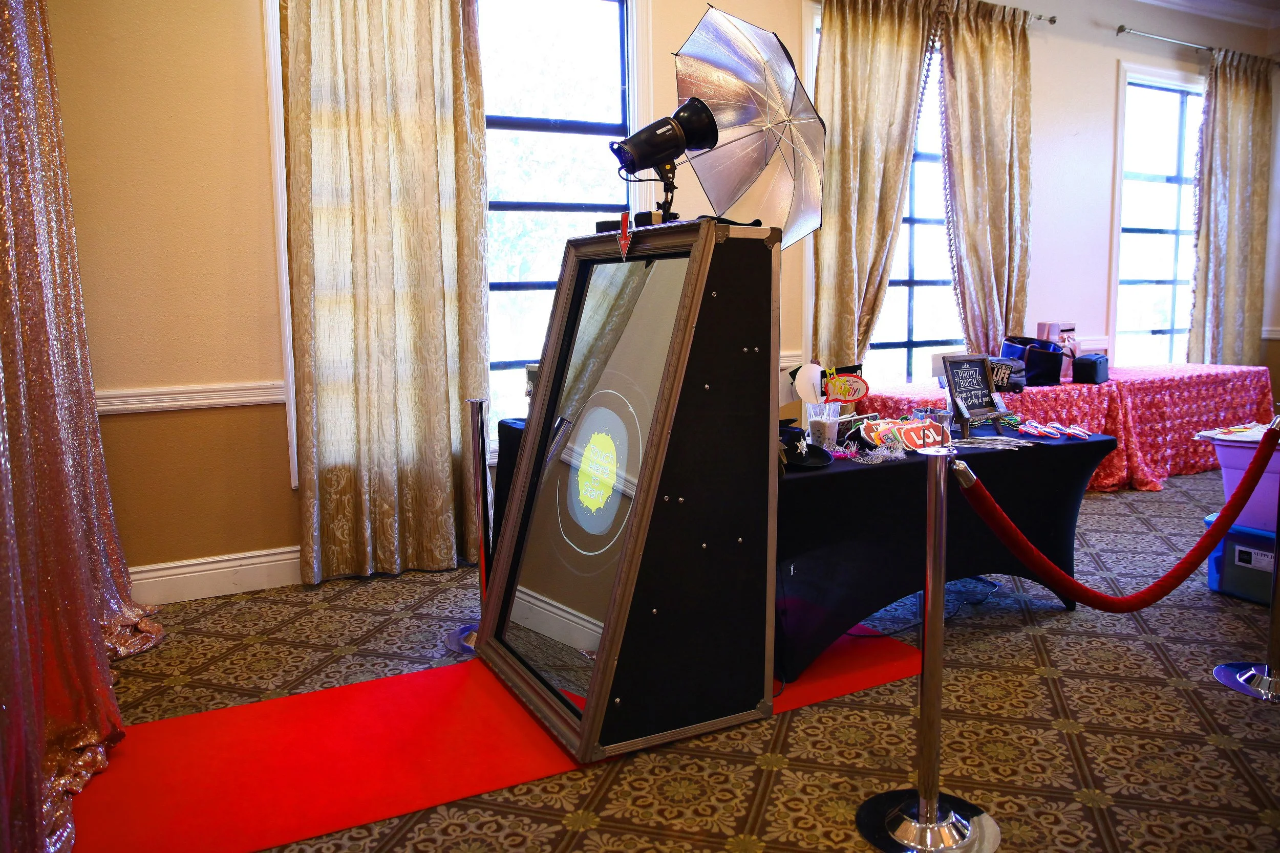 Photo booth with a large mirror, umbrella, and studio light, set up in a room with gold curtains and patterned carpet.