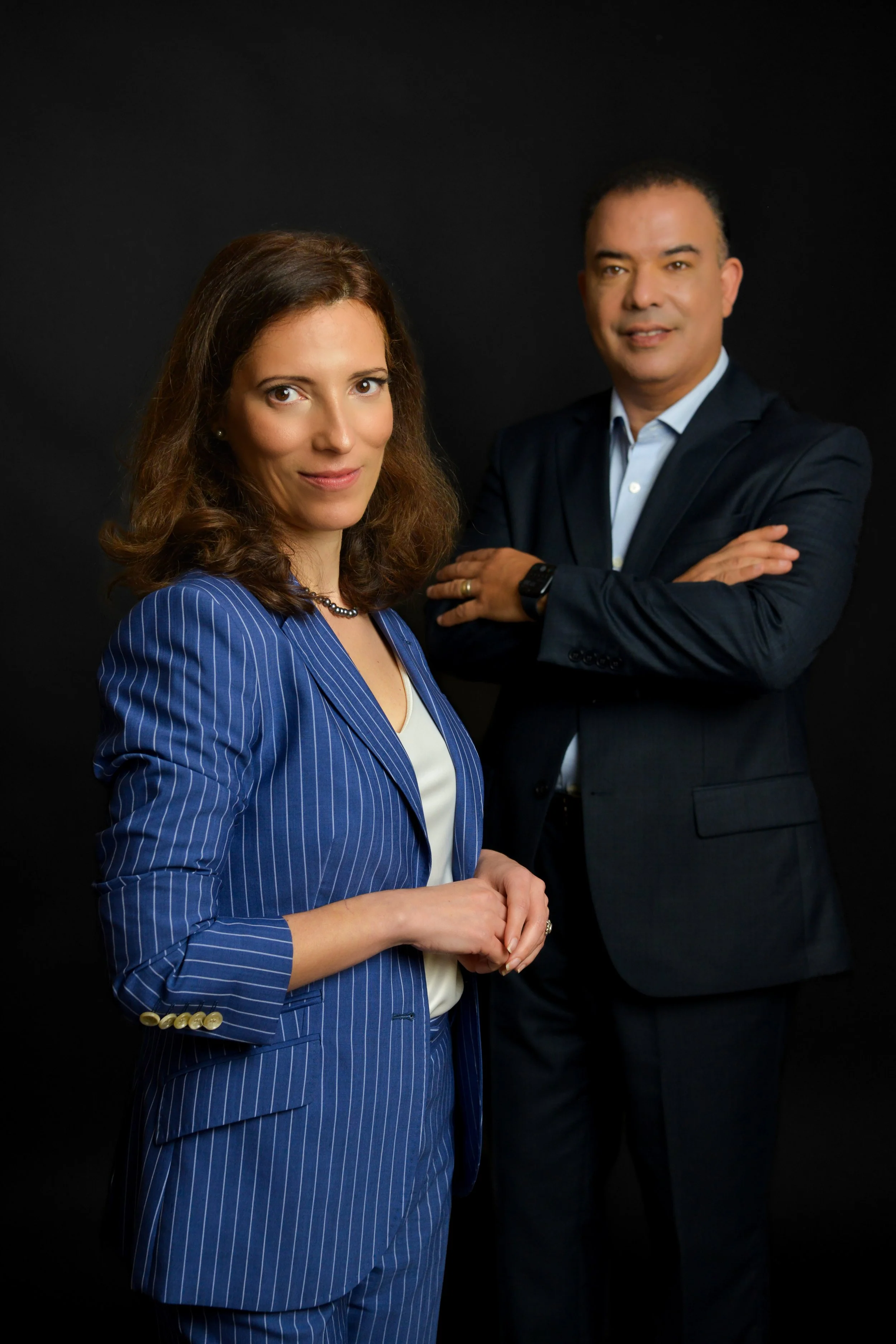 A professional woman with shoulder-length brown hair, wearing a blue pinstripe suit and a white blouse, standing with her hands clasped in front of her. A man with short dark hair, wearing a dark suit and a light blue shirt, is standing behind her with crossed arms. Both are against a black background.