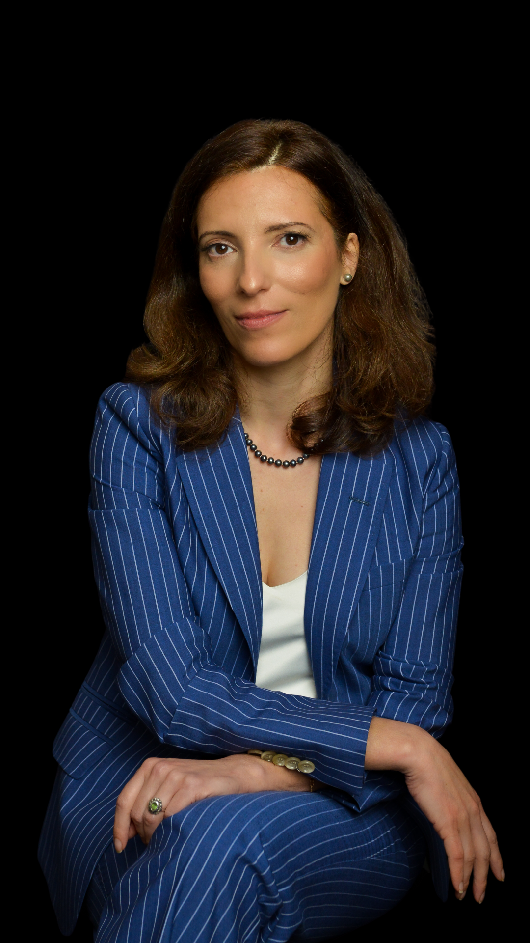 A woman with brown hair, wearing a blue pinstripe suit, white top, pearl earrings, and a pearl necklace, sitting with her arms crossed against a black background.