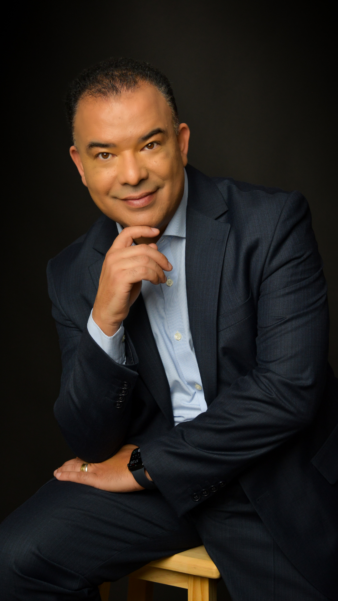 Man in a dark suit sitting on a wooden stool against a black background, looking directly at the camera with his right hand touching his chin.