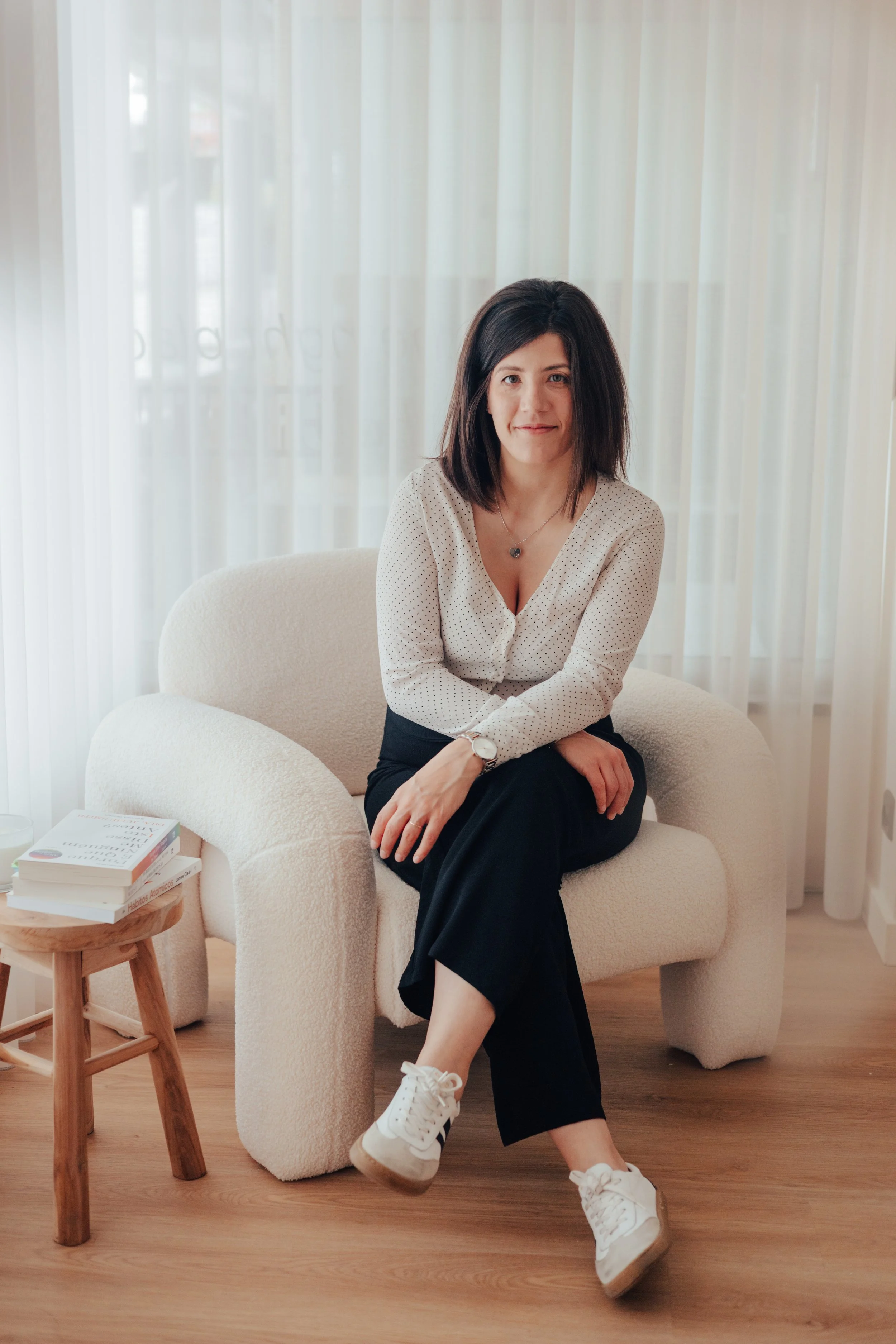 Mulher sentada em uma poltrona de cor creme, com cenário de cortinas brancas ao fundo, usando blusa branca com bolinhas pretas, calça preta, tênis branco e acessório de colar, sorrindo para a câmera.
