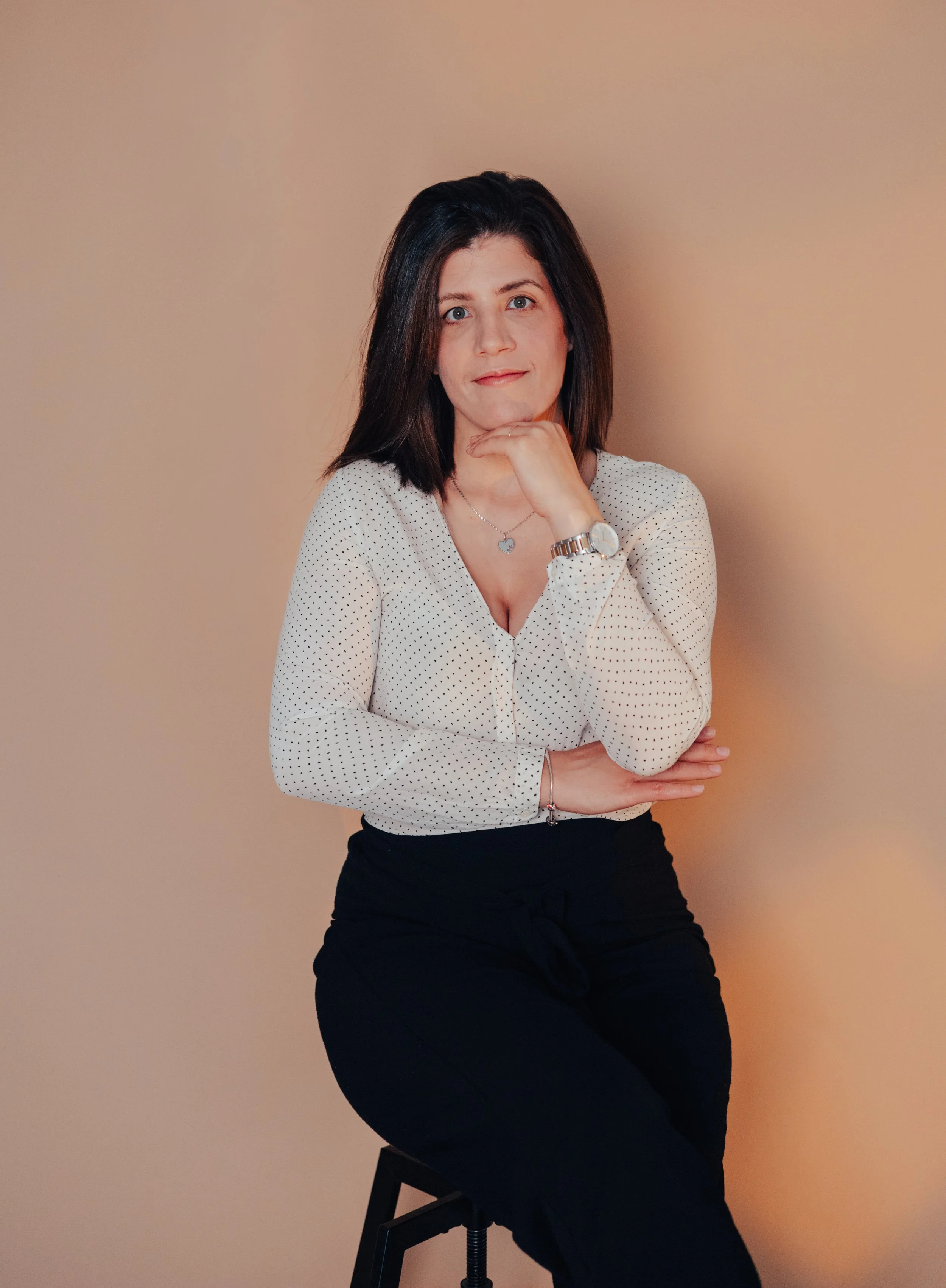 Mulher com cabelo castanho escuro, usando blusa de mangas compridas branca com pintas pretas e calça preta, sentada em uma cadeira preta, com o braço direito cruzado sobre a perna e a mão apoiada no queixo, contra fundo bege.