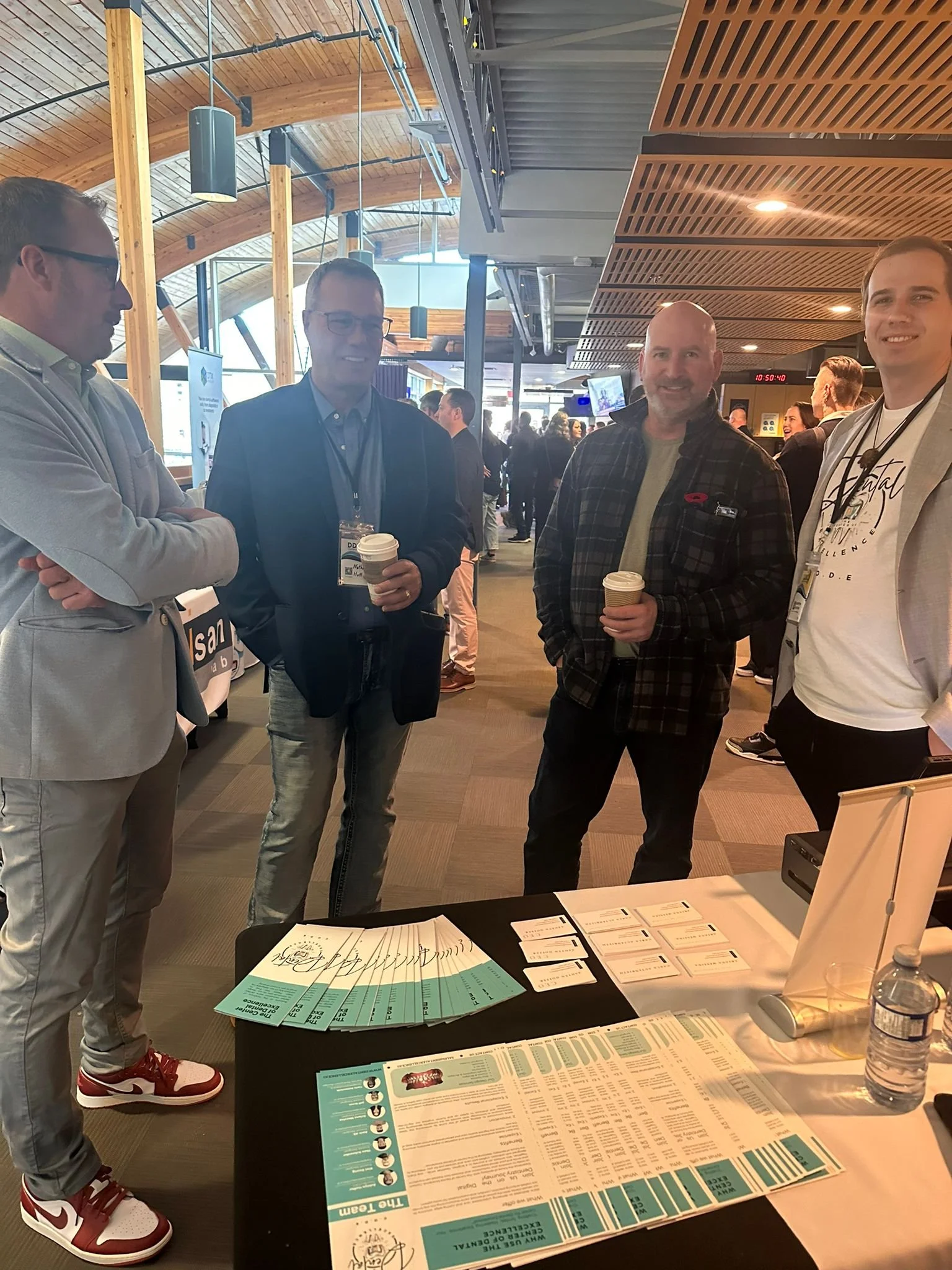 Four men standing at a conference booth, holding coffee cups, with informational brochures and tickets on the table in front of them. The background shows a modern, well-lit conference venue with other attendees.