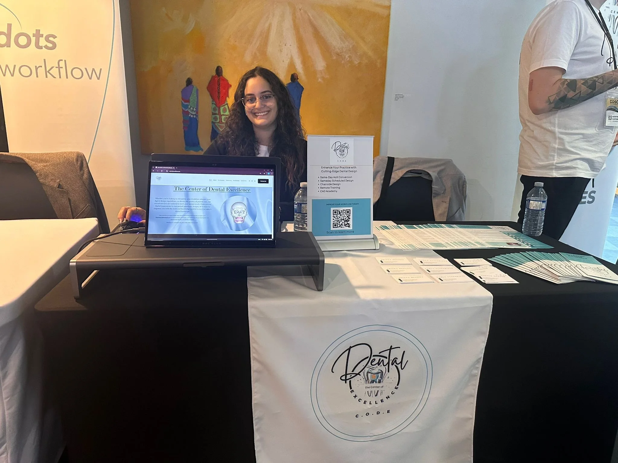 A woman with glasses and long curly hair sitting behind a display table at a dental conference booth, smiling at the camera. The table has brochures, business cards, and a sign with a QR code. There is a laptop on the table showing a website about de