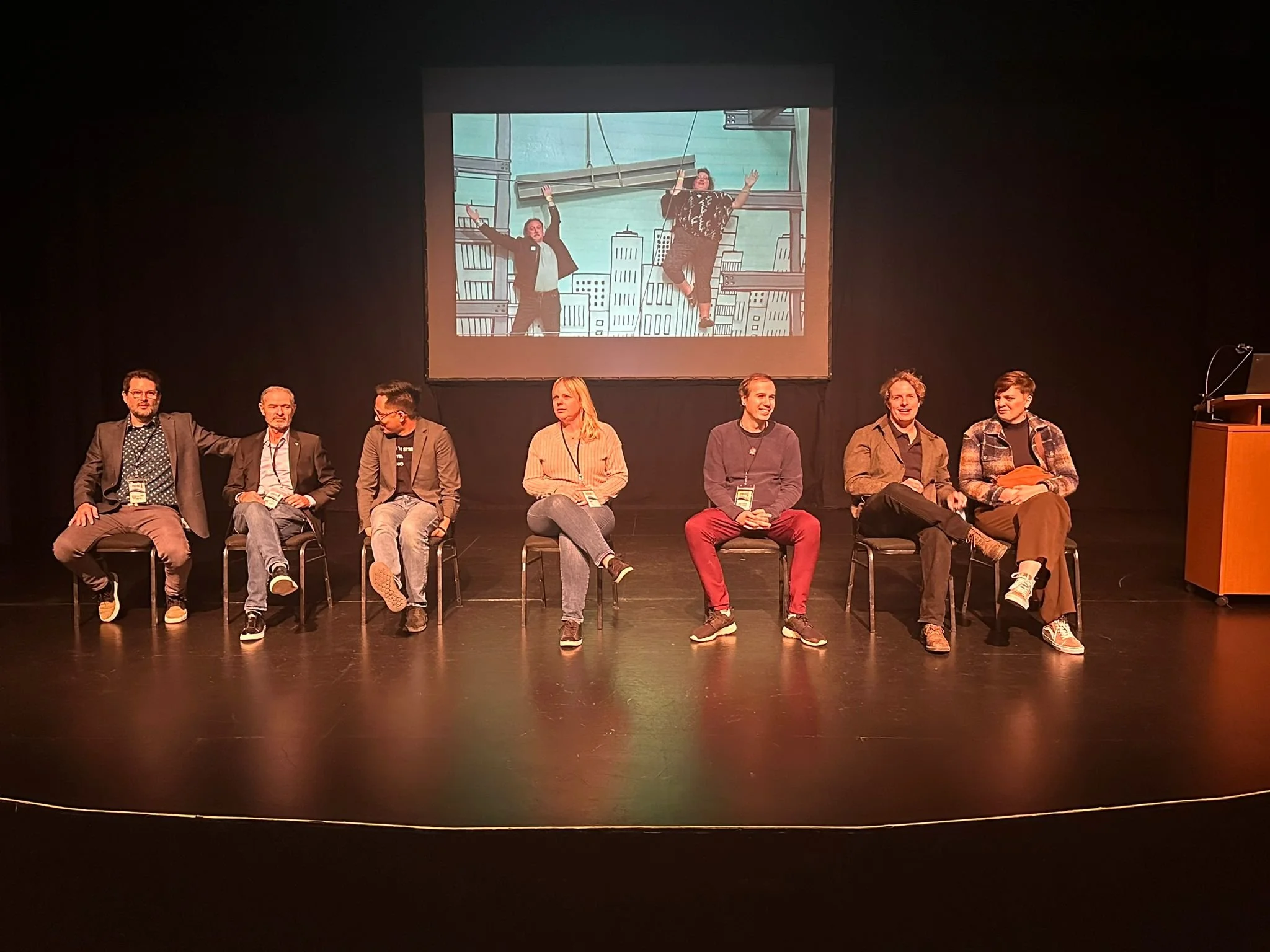A panel of seven people sitting on chairs on a stage, with a large animated background projection of two characters in a cityscape behind them.