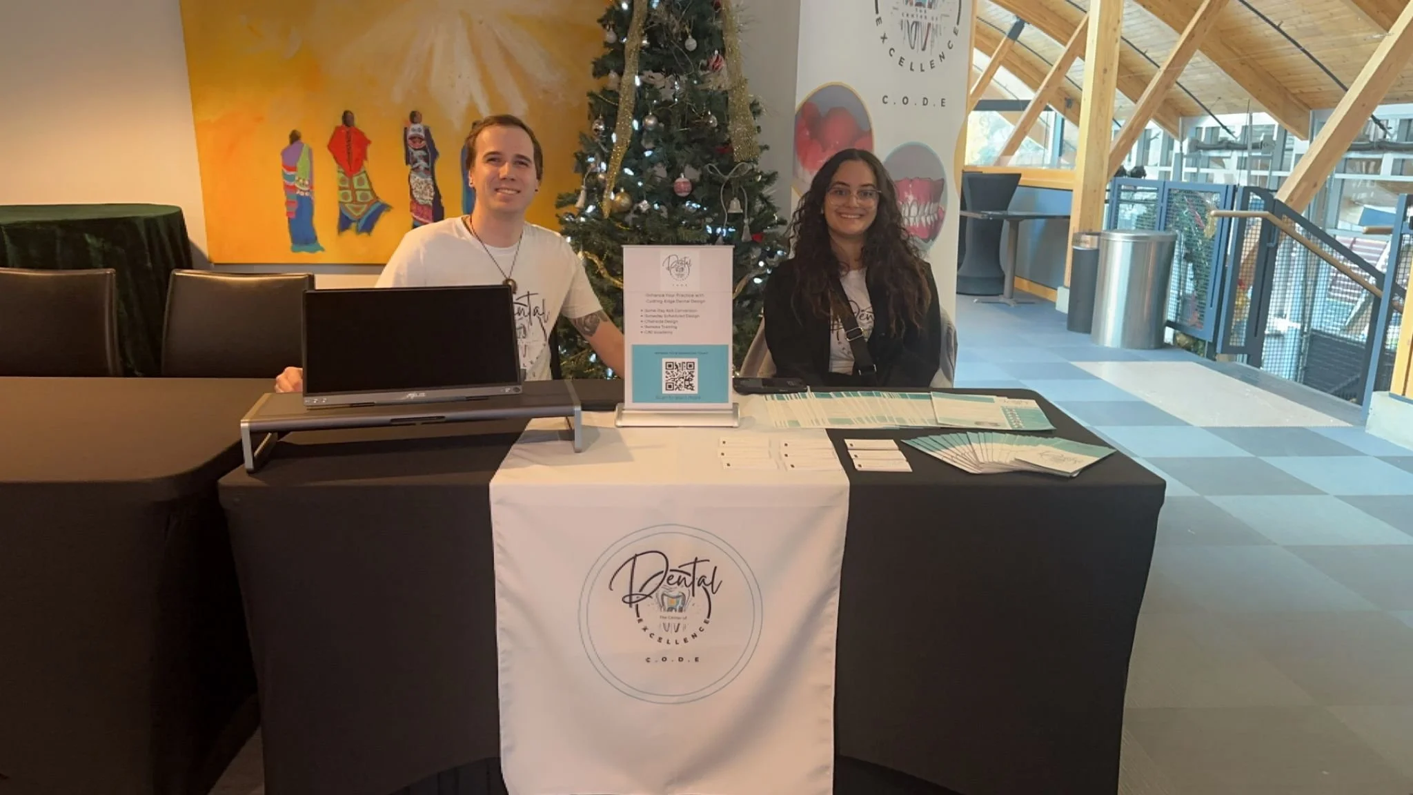 Two smiling people sitting behind a table at a dental event with a Christmas tree and colorful artwork in the background.