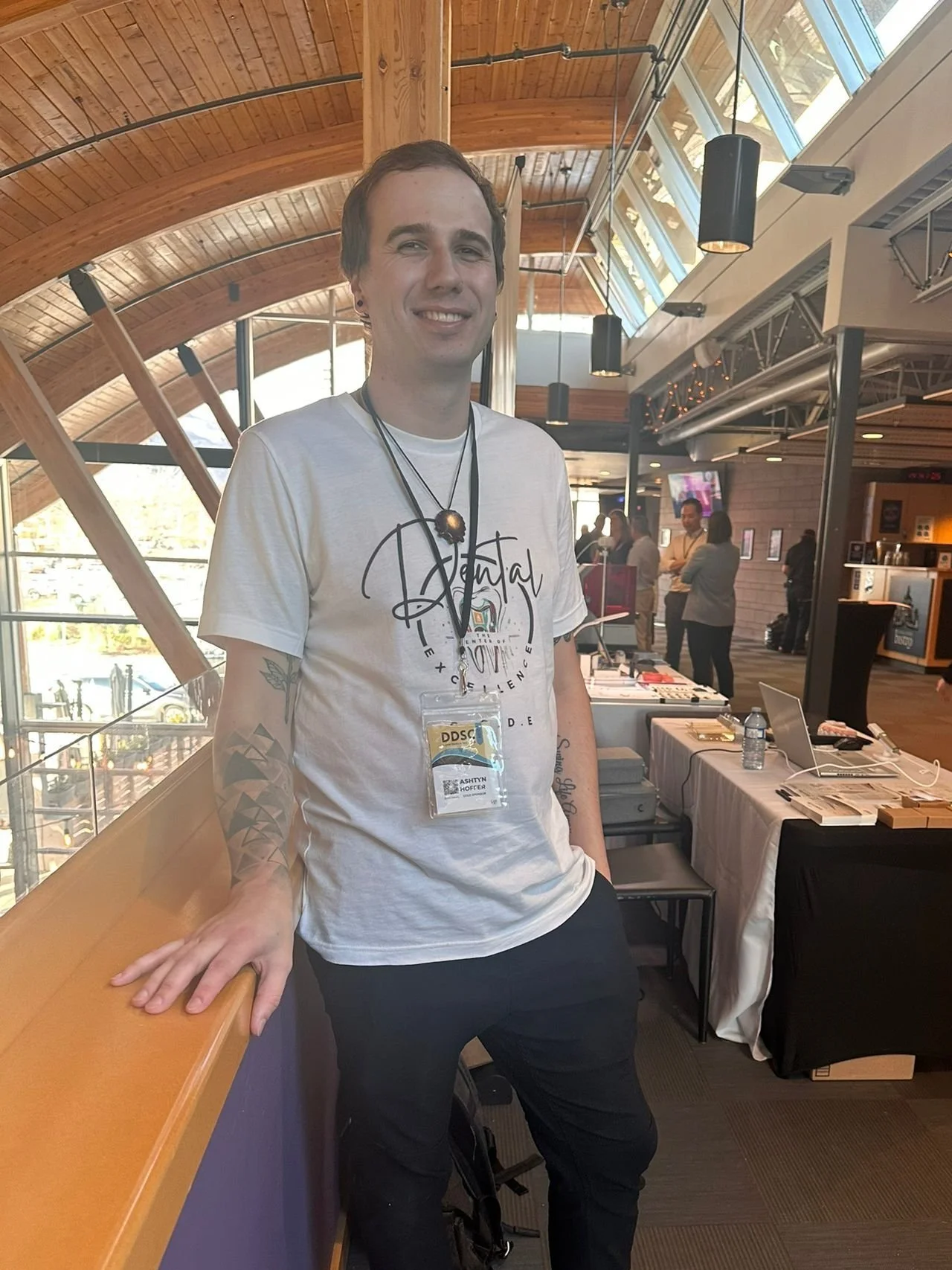 A young man with tattoos on his arms smiling and standing in a conference hall with wooden and glass architecture. He is wearing a white t-shirt, a name badge, and two necklaces.