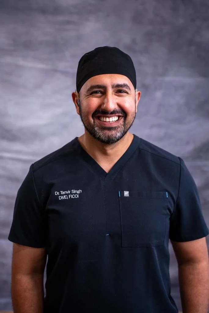 A man in black medical scrubs and a black head covering smiling in front of a gray textured background.