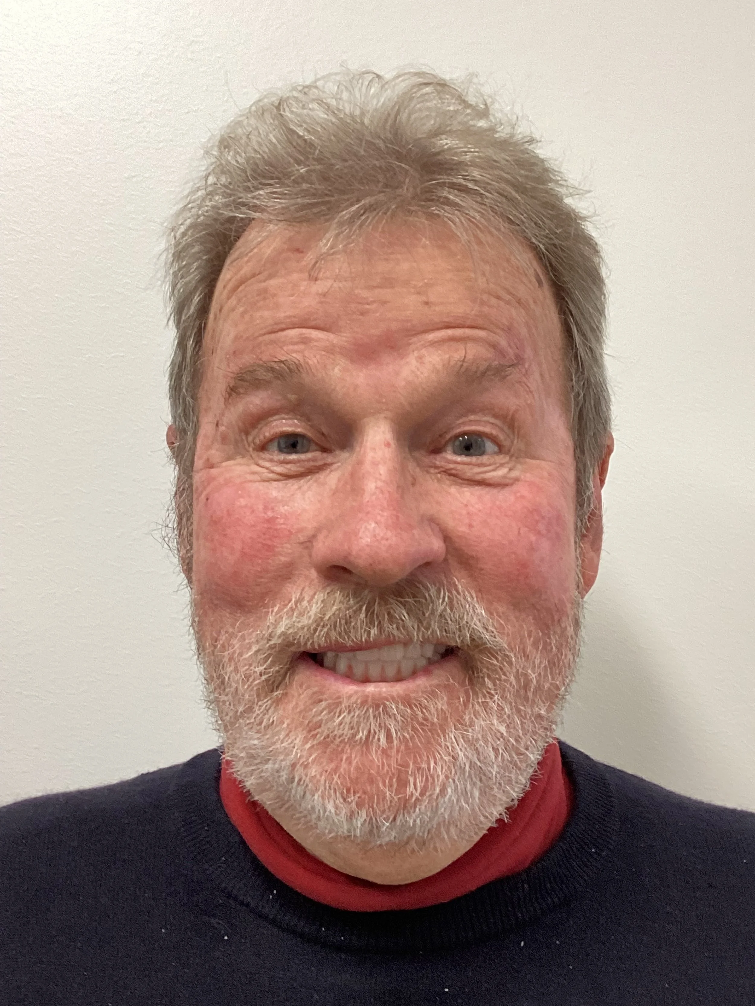 A middle-aged man with gray hair and a beard smiling in front of a plain white wall.