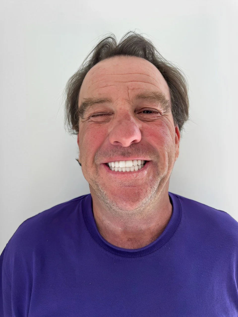 A middle-aged man with light skin, brown hair, and a beard, smiling widely with one eye partially closed, wearing a purple shirt against a plain white background.