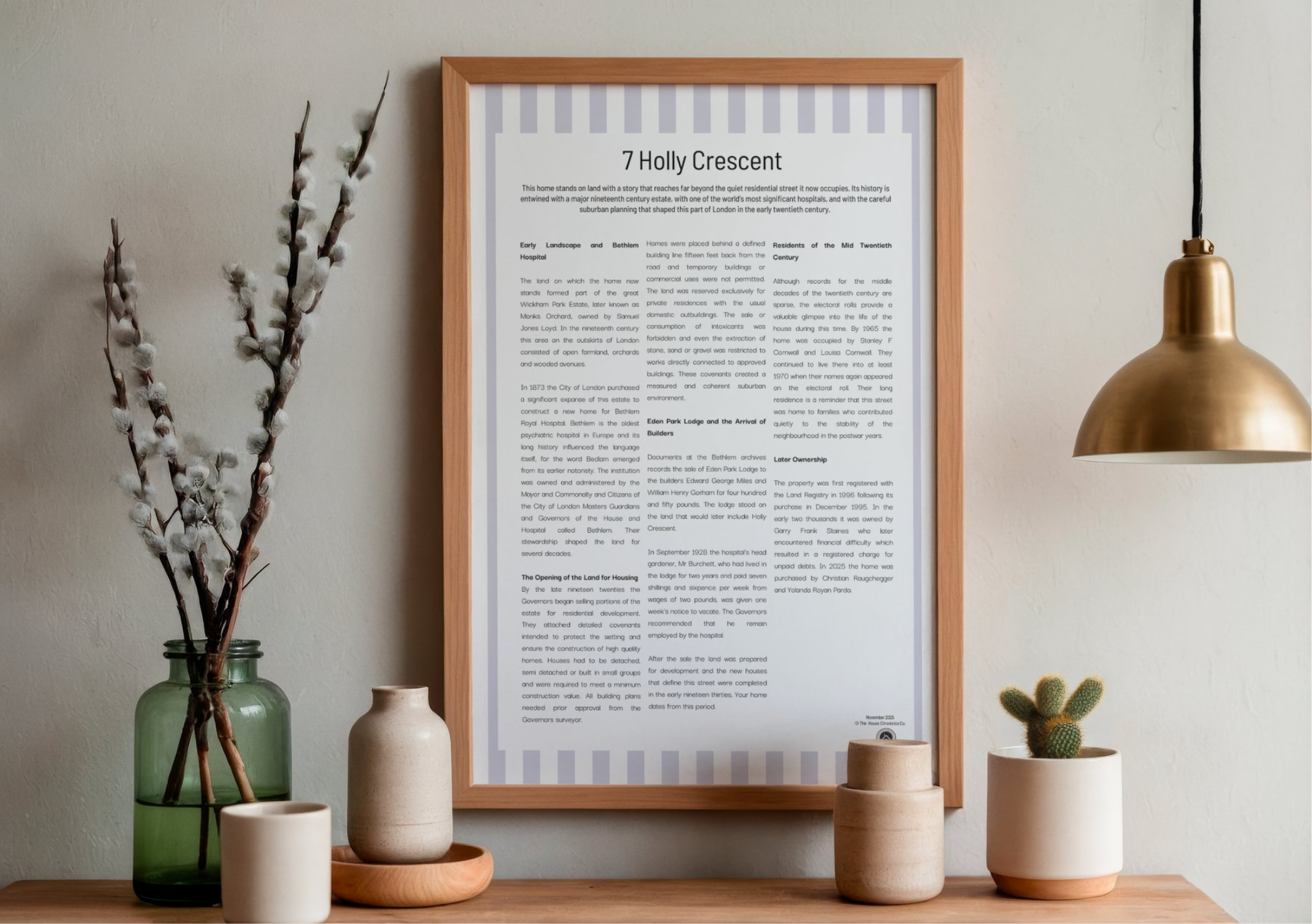 Decorative interior with a framed informational poster titled '7 Holly Crescent', a green glass vase with cotton branches, a small beige vase on a wooden tray, a white cylindrical vase, a cactus in a white pot, and a gold desk lamp hanging from the ceiling on a wooden surface against a beige wall.