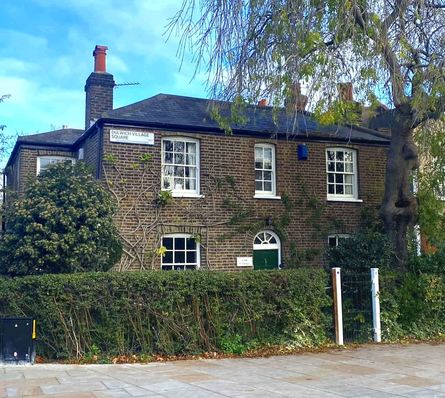 Brick house with white window frames, ivy and bushes in front, a sign indicating Dulwich Village Square, and a metal gate in a garden, with a tree on the right side.