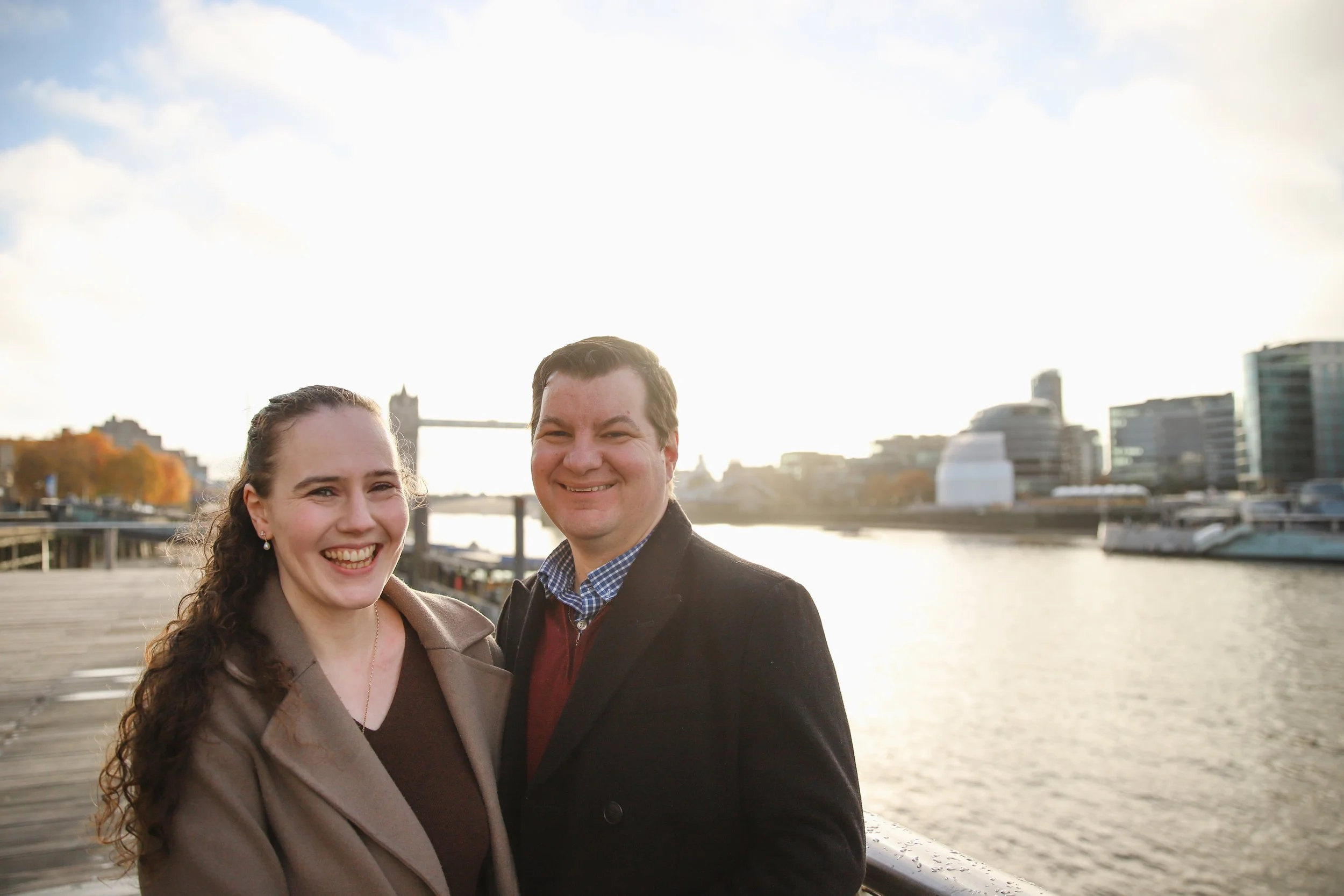 A smiling young woman and a young man standing together near the water with a city skyline and a bridge in the background during sunset.
