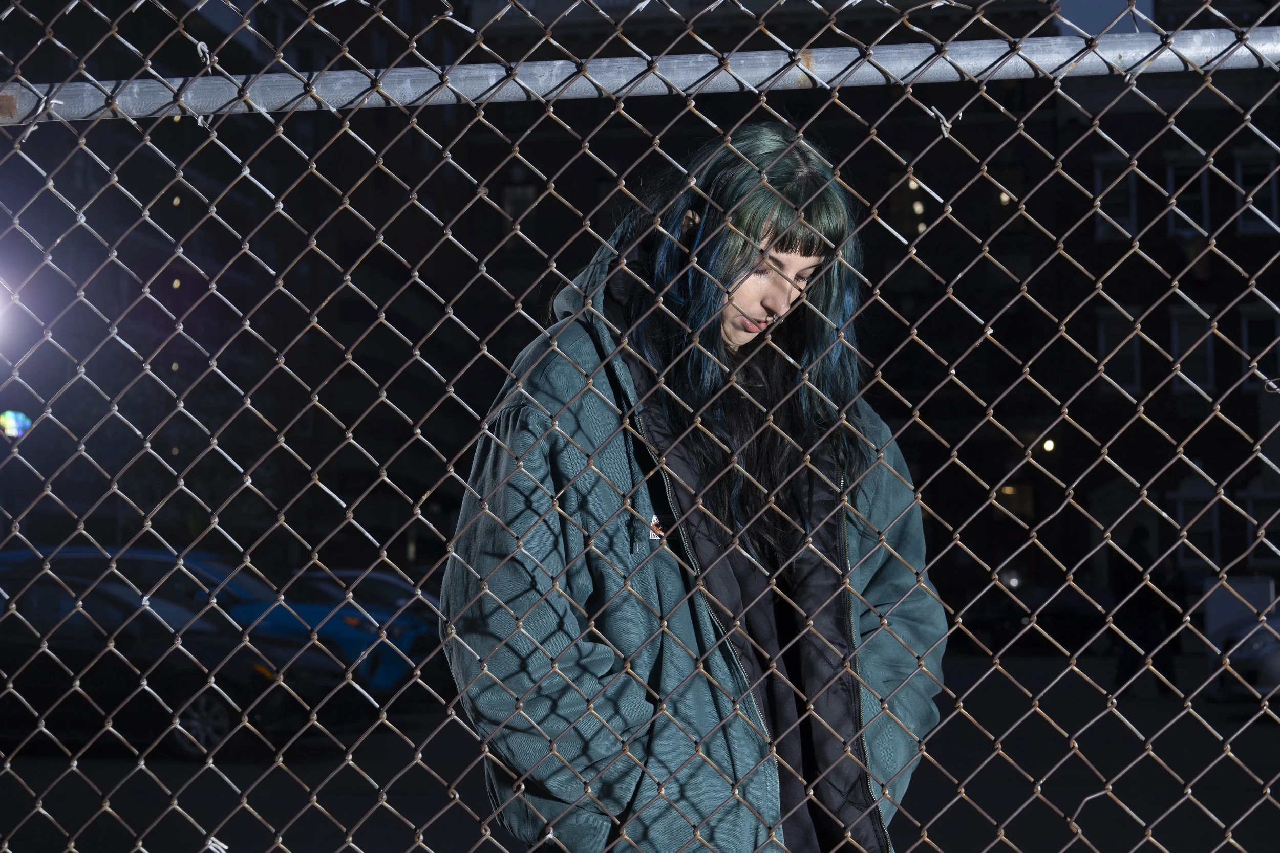 A person with dark hair and green highlights stands behind a chain-link fence at night, smiling with eyes closed, wearing a teal jacket and a black hoodie.