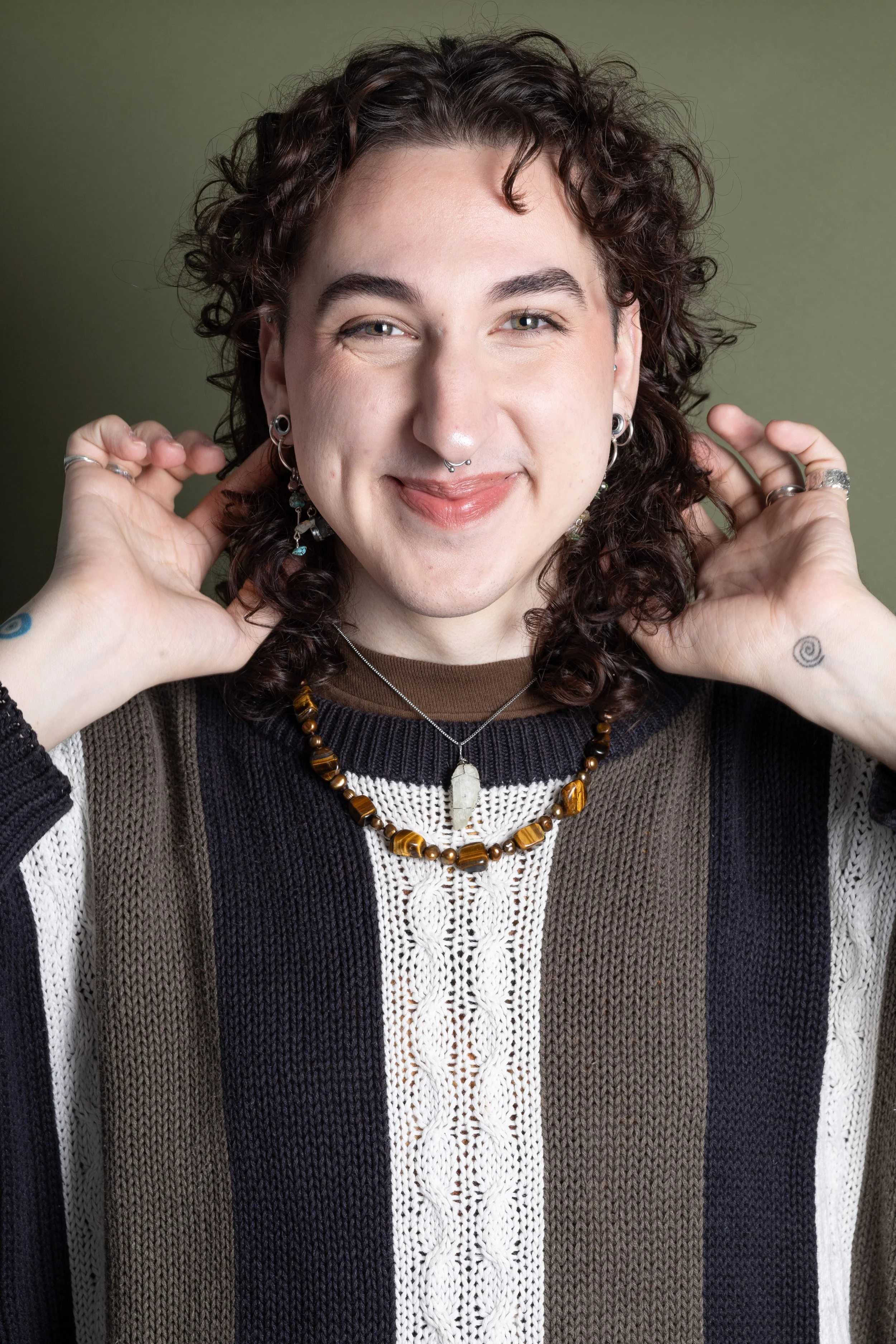 A person with curly brown hair smiling, wearing layered necklaces, earrings, and a striped sweater, with hands near their ears.