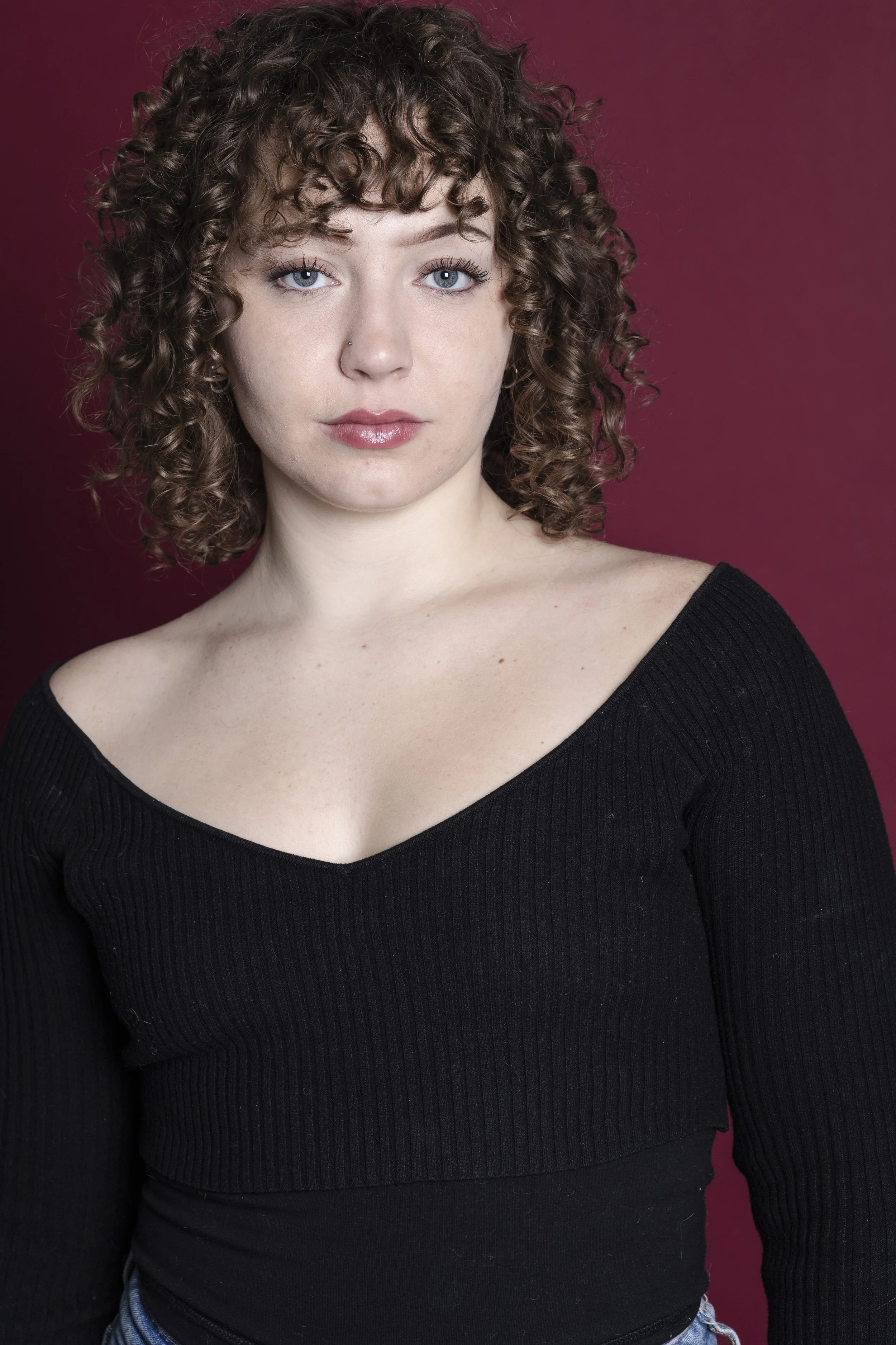 Portrait of a young woman with curly brown hair, wearing a black top, against a maroon background.