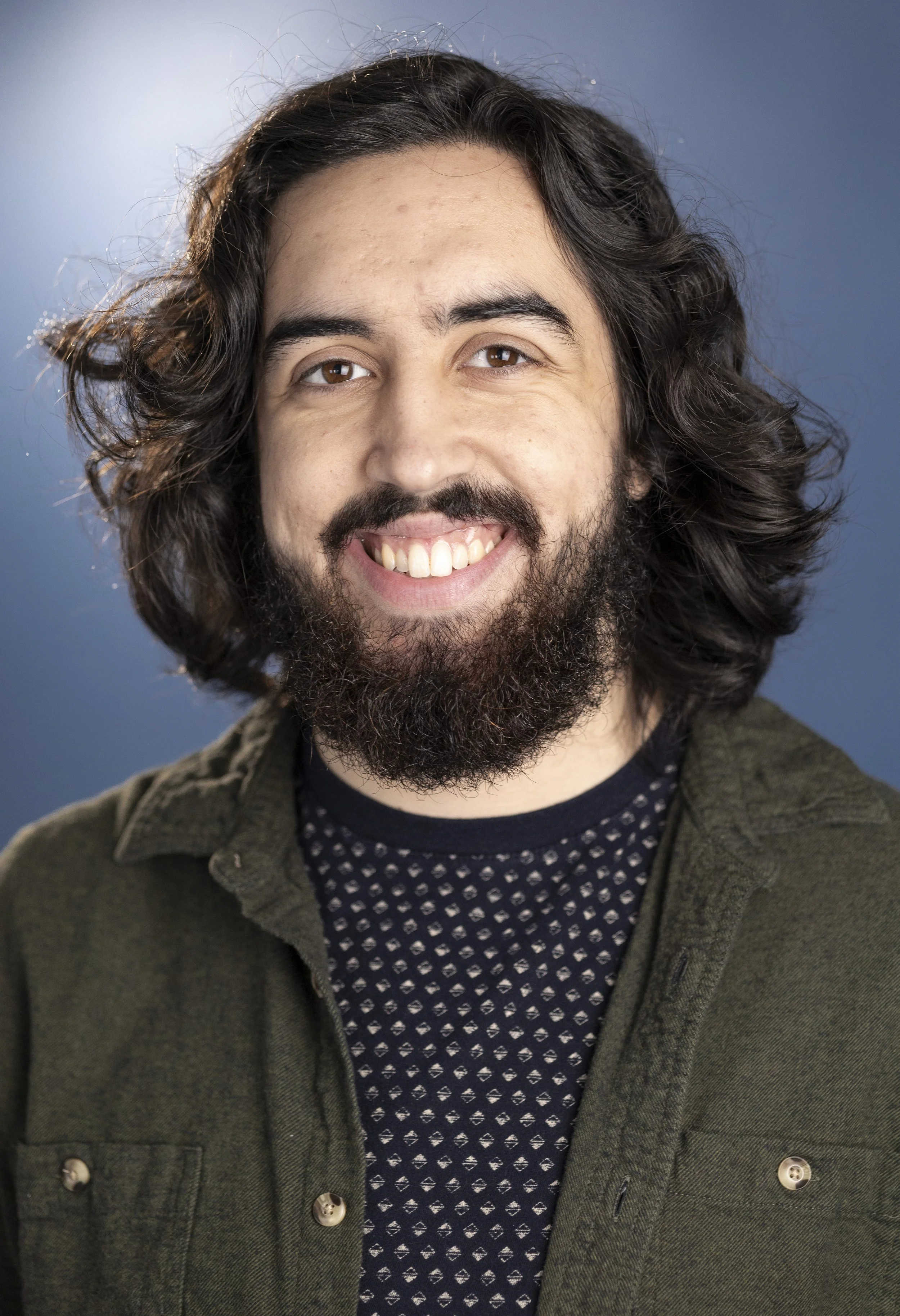 A smiling young man with long wavy dark hair, a full beard, and brown eyes, wearing a dark patterned shirt and an olive green jacket, against a blue background.
