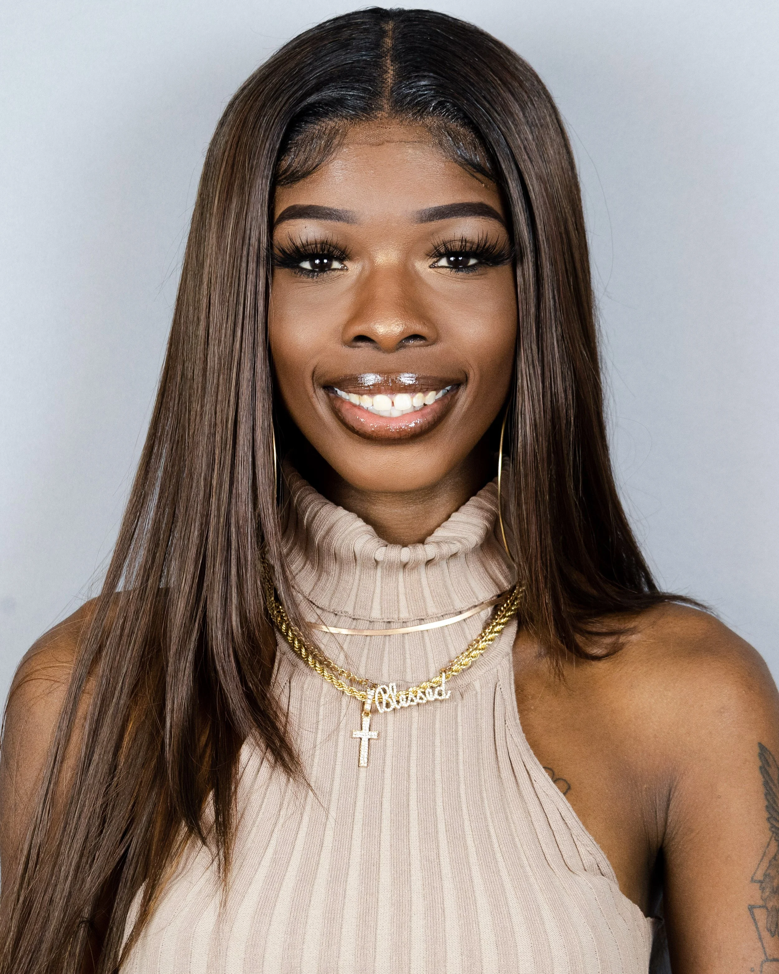 Portrait of a young woman with long brown hair, wearing a beige turtleneck top and gold jewelry, smiling at the camera.