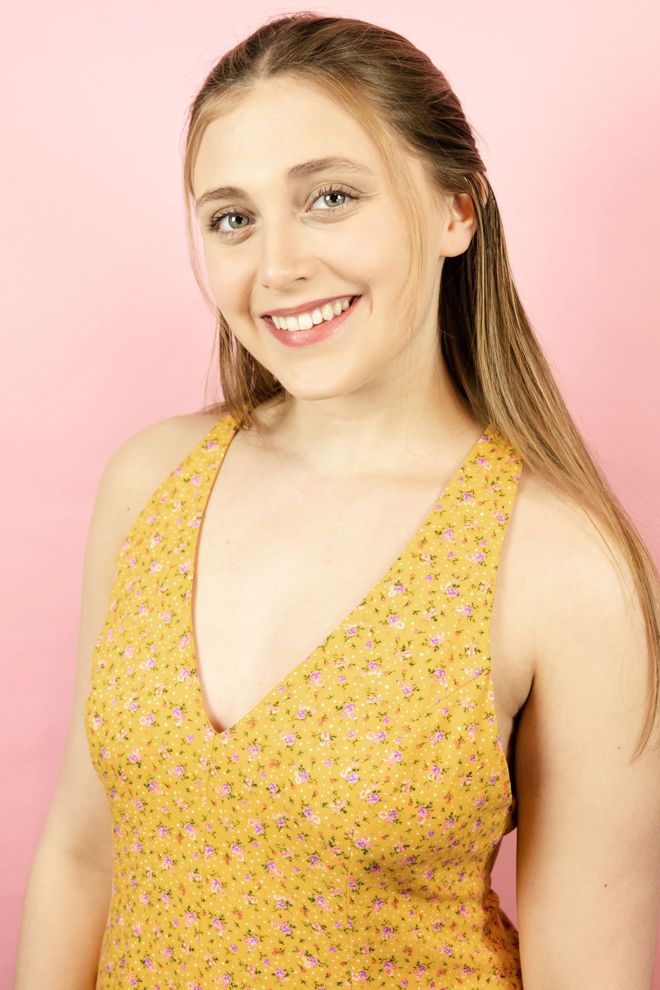 A young woman with long light brown hair, smiling, wearing a yellow floral dress, against a pink background.