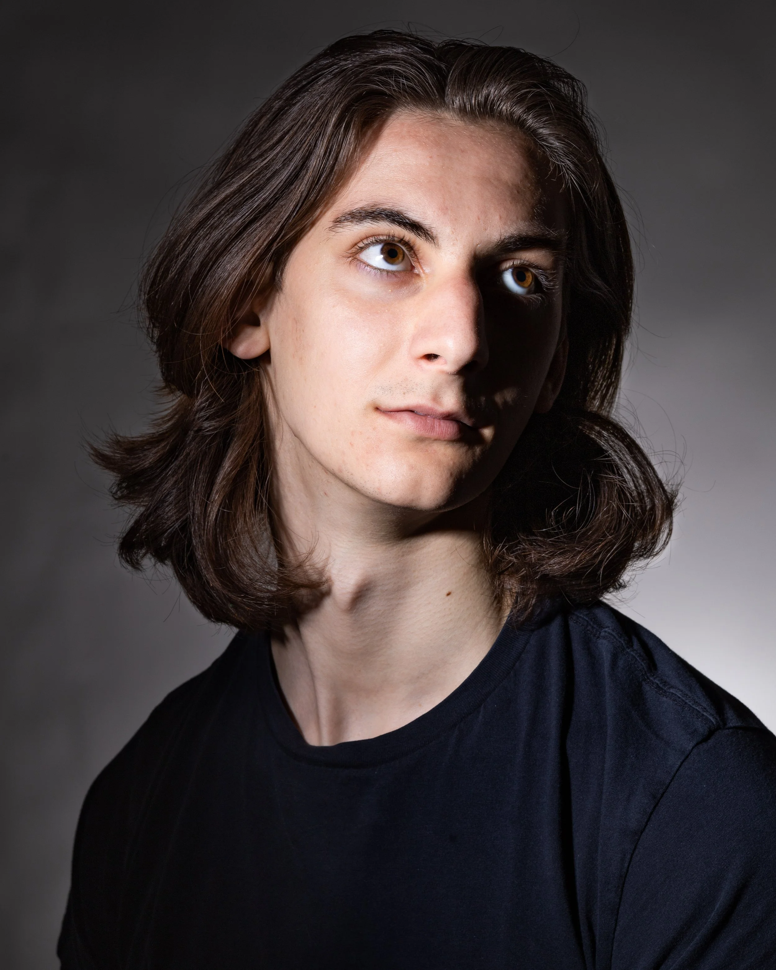 A young person with shoulder-length brown hair, wearing a black shirt, looking slightly to the side against a dark gradient background.