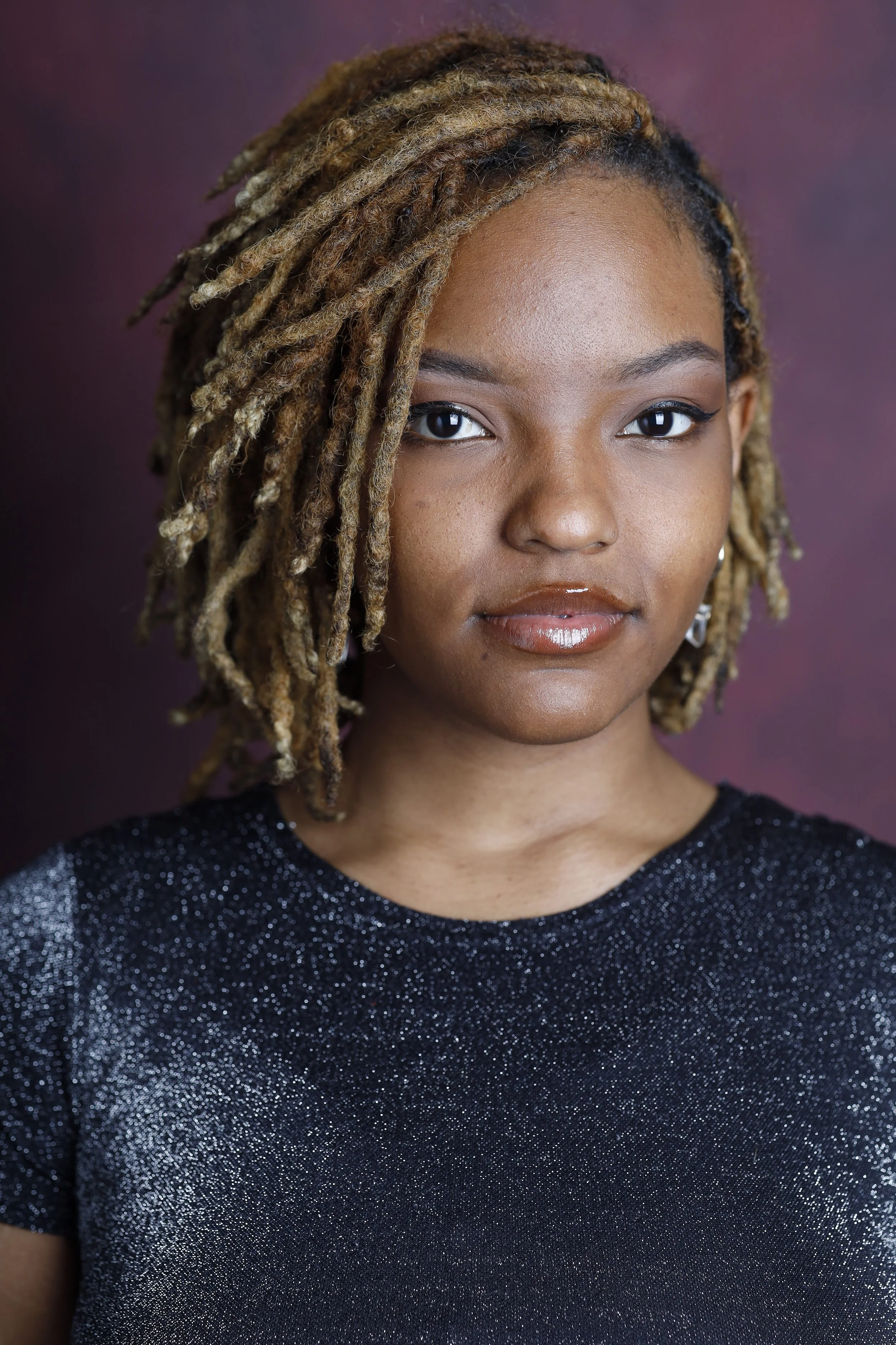 Portrait of a young woman with braided blonde hair, wearing a sparkly black top, looking directly at the camera against a dark purple background.